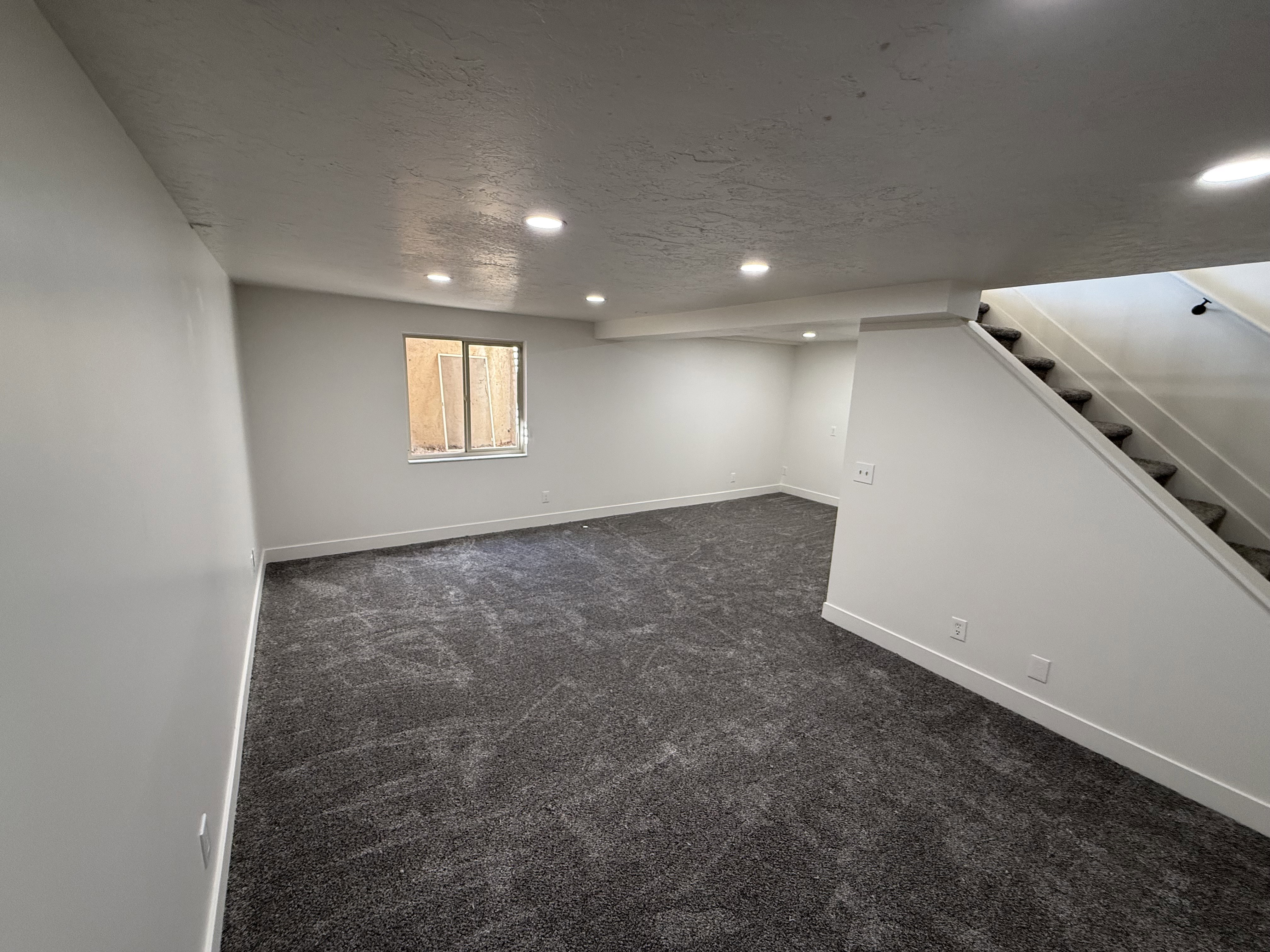 Spacious interior room featuring plush gray carpeting, recessed ceiling lighting, and a window providing natural light
