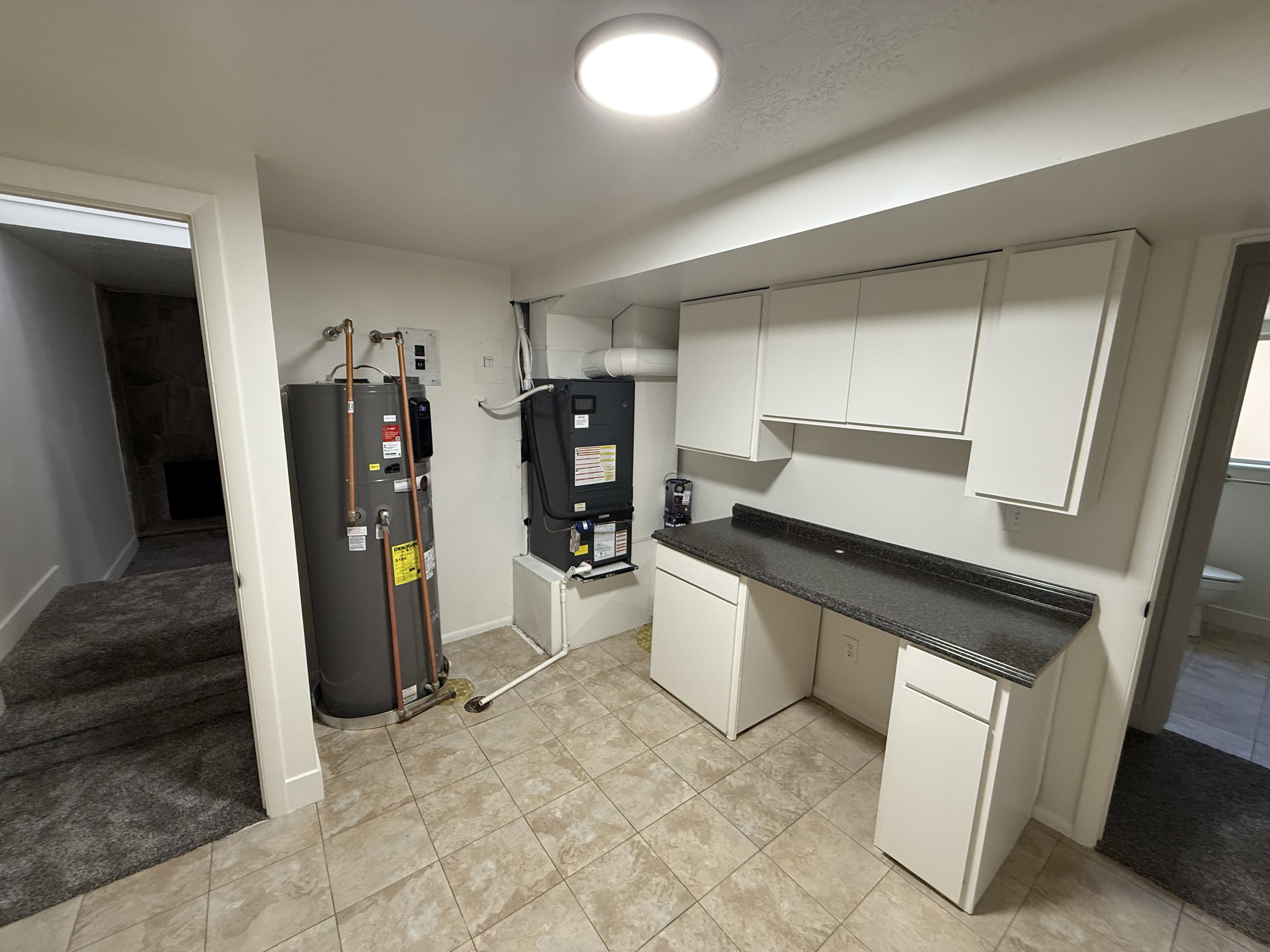 Utility space featuring light-colored tile flooring, white cabinetry, dark countertop, and exposed water heater