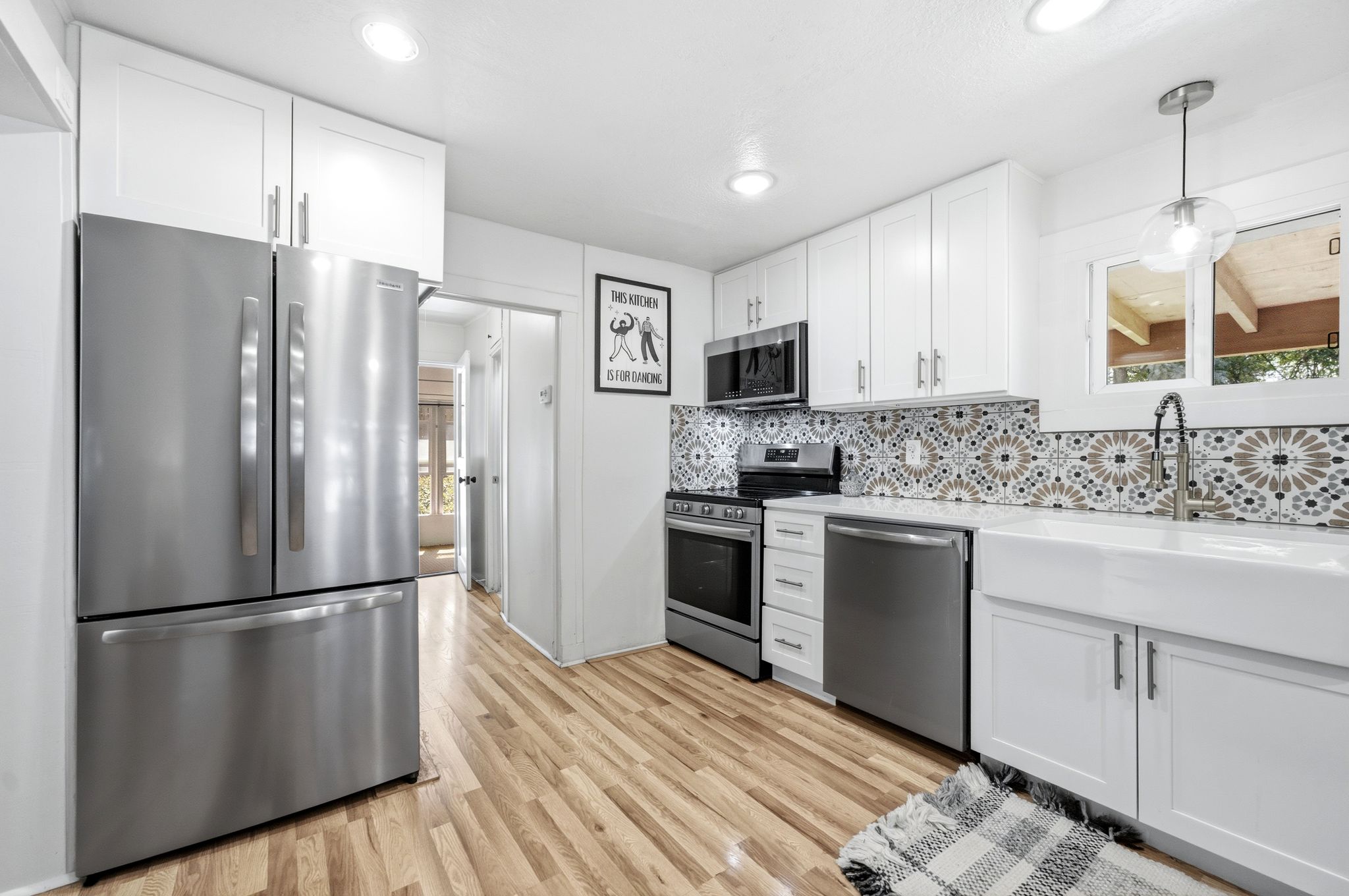 Kitchen featuring stainless steel appliances, patterned tile backsplash, white shaker cabinetry, wood-finish flooring, and a farmhouse sink