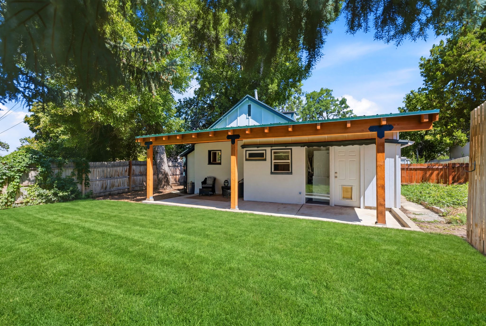 Expansive backyard featuring a manicured lawn, a covered patio with exposed wood beams, and a white stucco exterior with a teal metal roof accent
