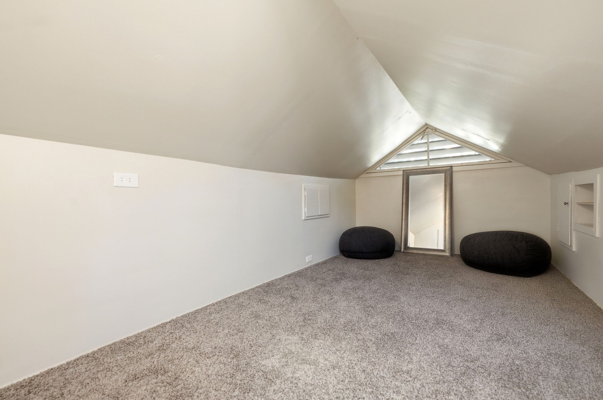 Carpeted attic space featuring sloped ceilings, a triangular louvered window, and built-in storage nooks