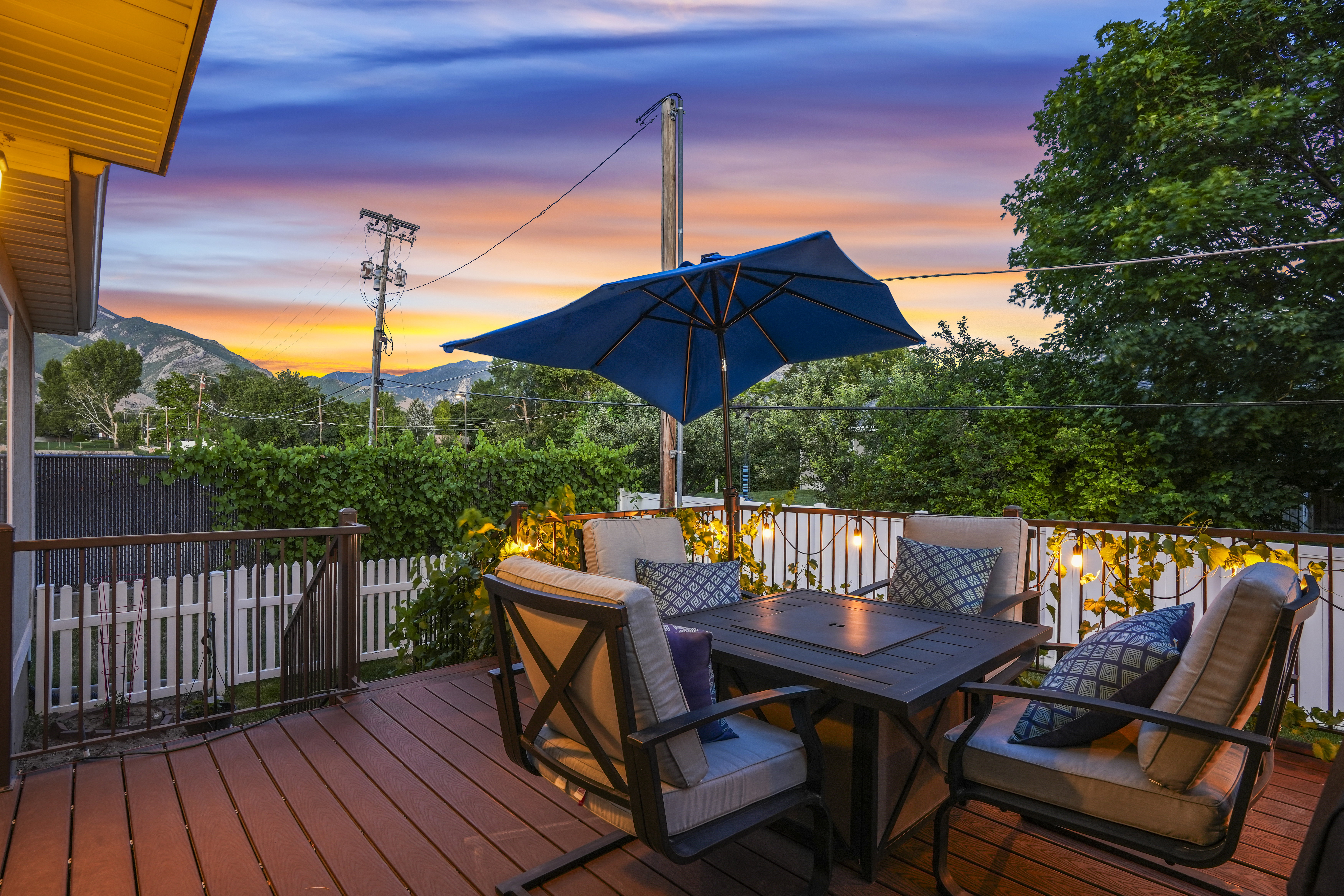 Expansive wood-finish deck featuring a built-in fire table, white picket fencing, and string lighting