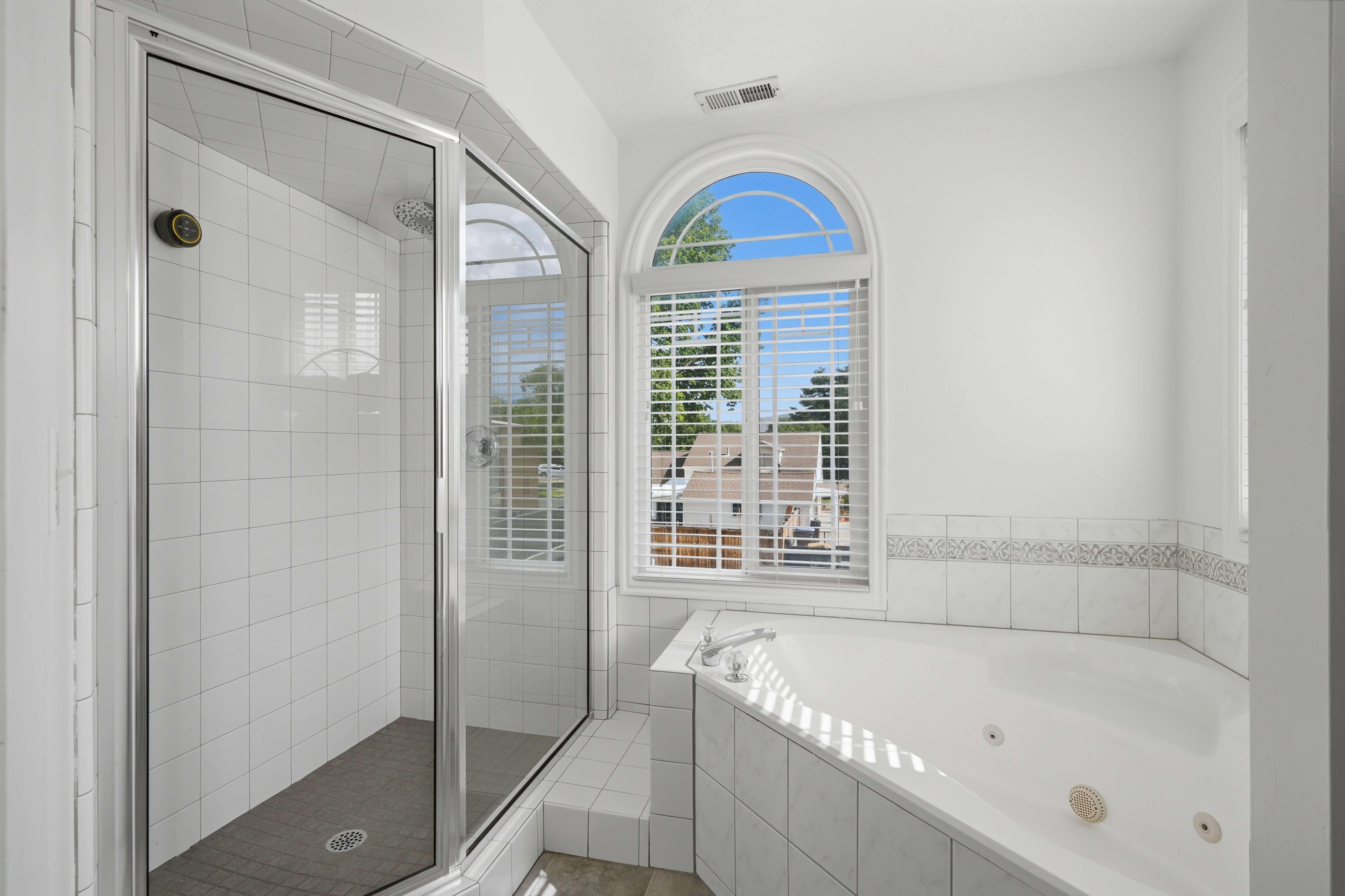 Spacious bathroom featuring a glass-enclosed shower with white subway tile, a built-in jetted tub with a tiled surround, and an arched window with blinds