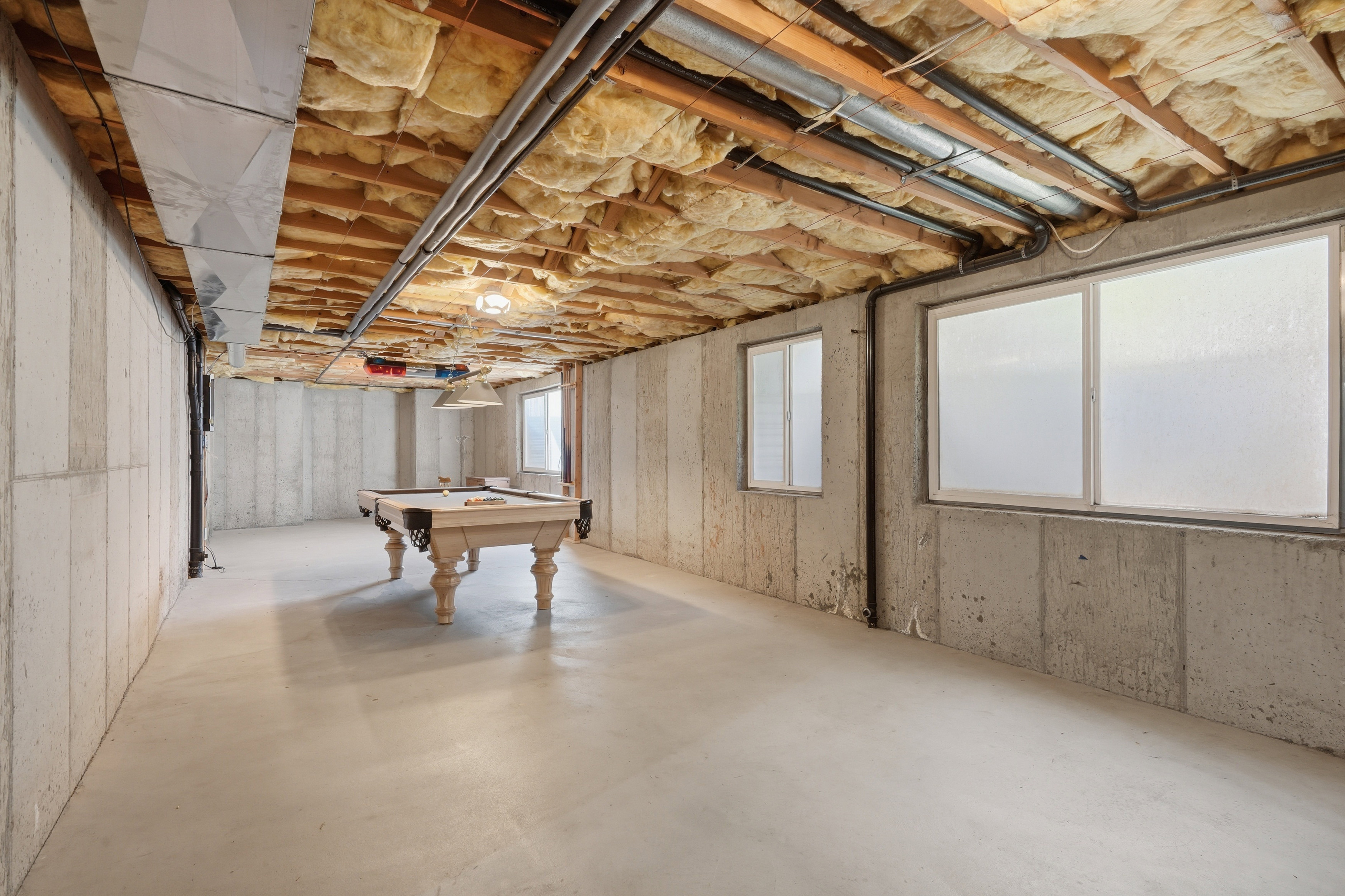 Spacious unfinished basement featuring concrete walls and flooring, exposed ceiling joists with insulation, two windows, and exposed ductwork