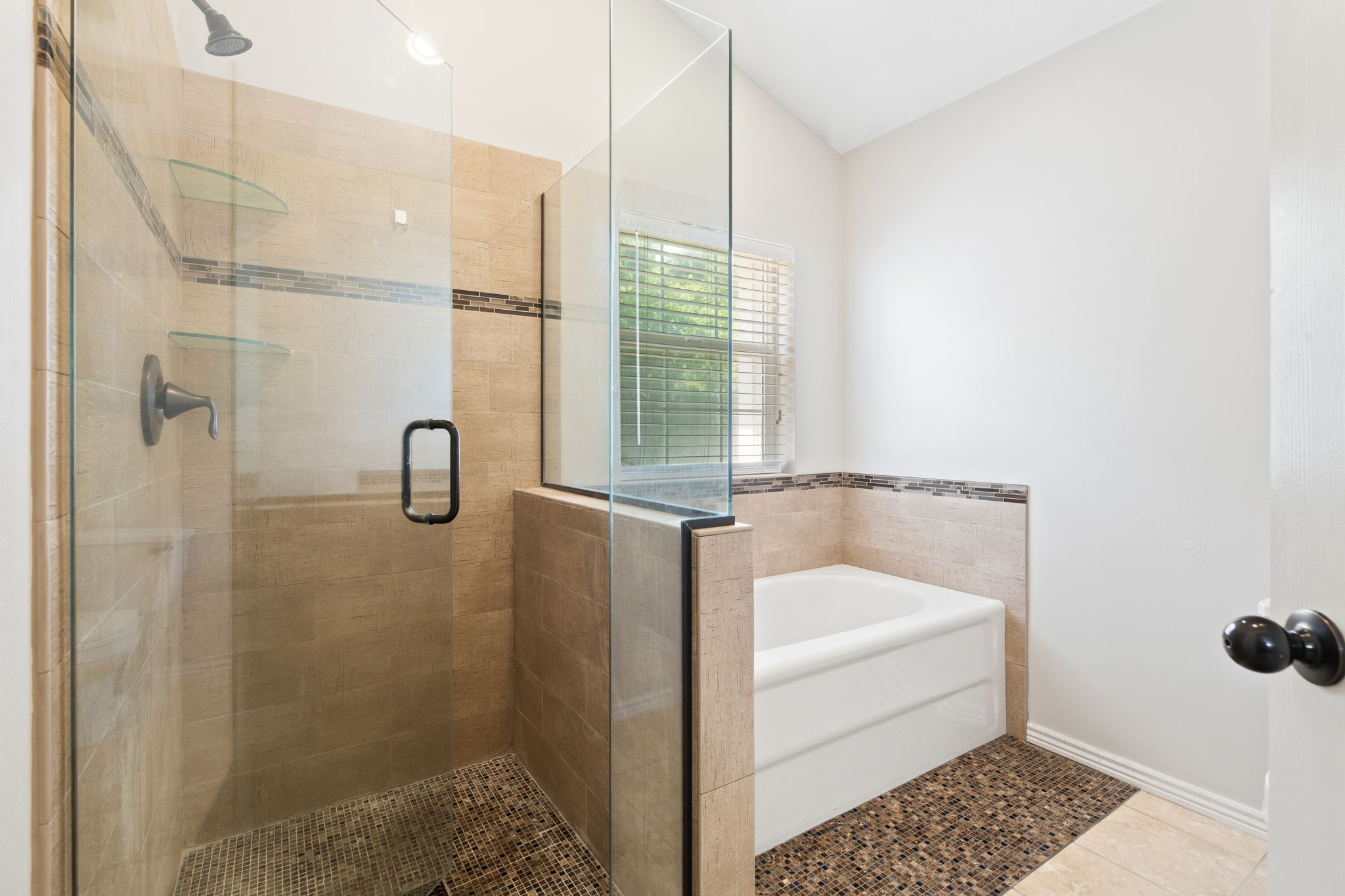 Bathroom featuring a glass-enclosed shower with tiled walls and two glass shelves, a white soaking tub, and a window with blinds