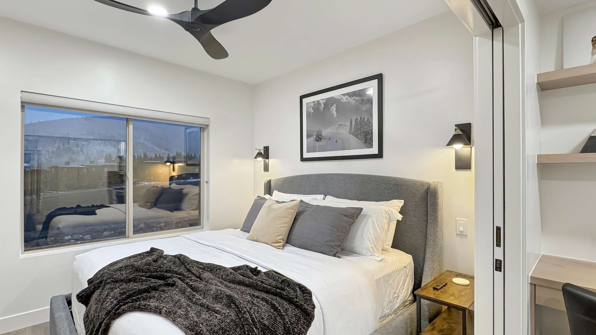Sleek bedroom featuring a modern ceiling fan, large sliding window, wall-mounted sconces, recessed lighting, and built-in shelving with an integrated desk