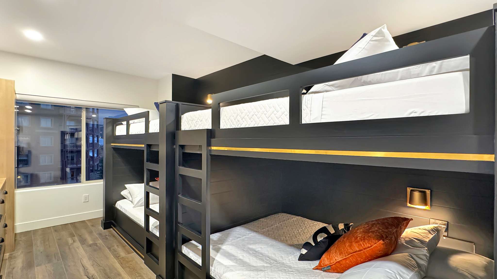Contemporary bedroom featuring built-in bunk beds with dark wood finish, integrated lighting, and a window with urban surroundings