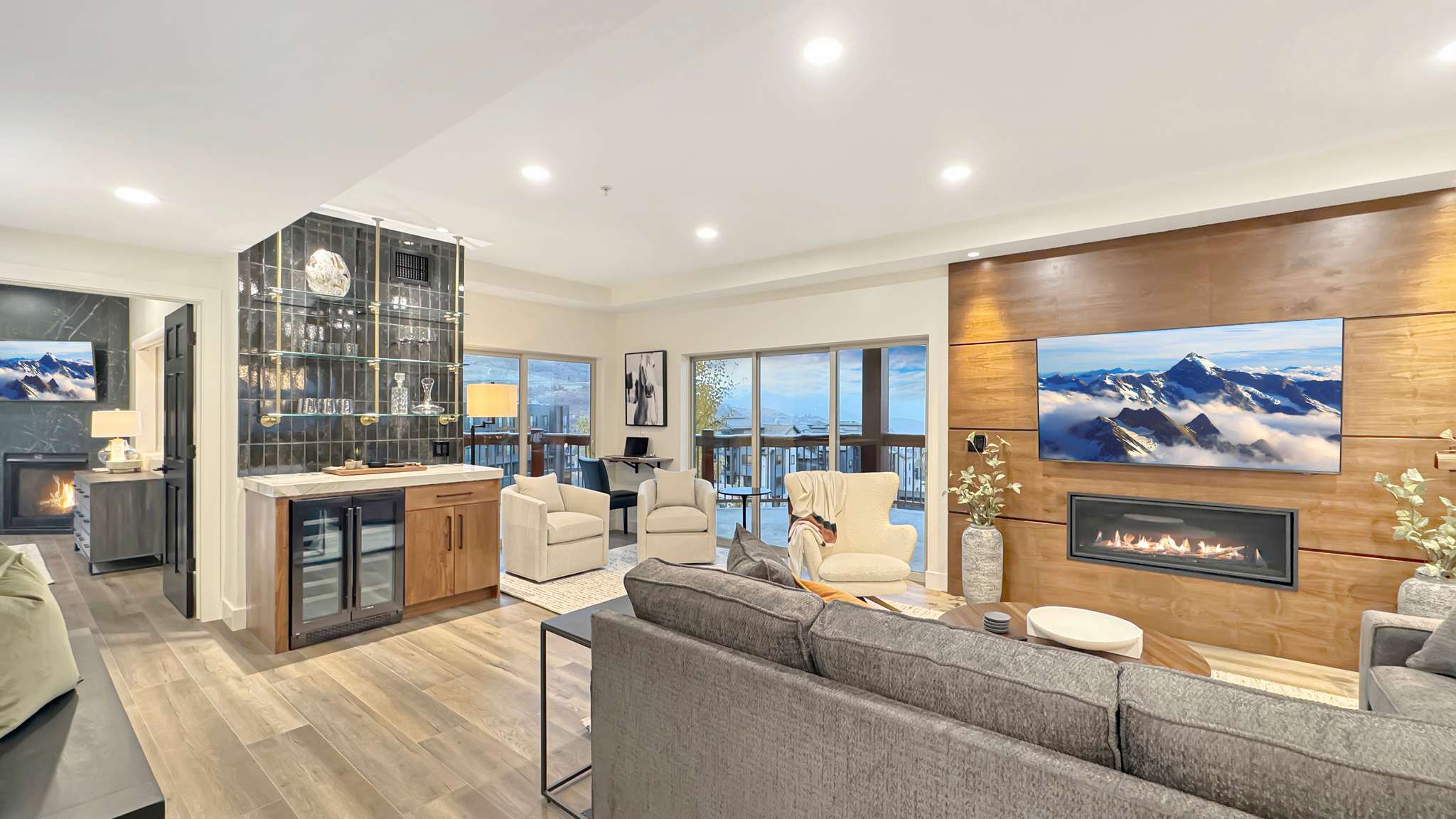 Spacious living area featuring wood-finish flooring, a linear electric fireplace set into a wood-paneled wall, a built-in wet bar with beverage coolers, and extensive glass doors