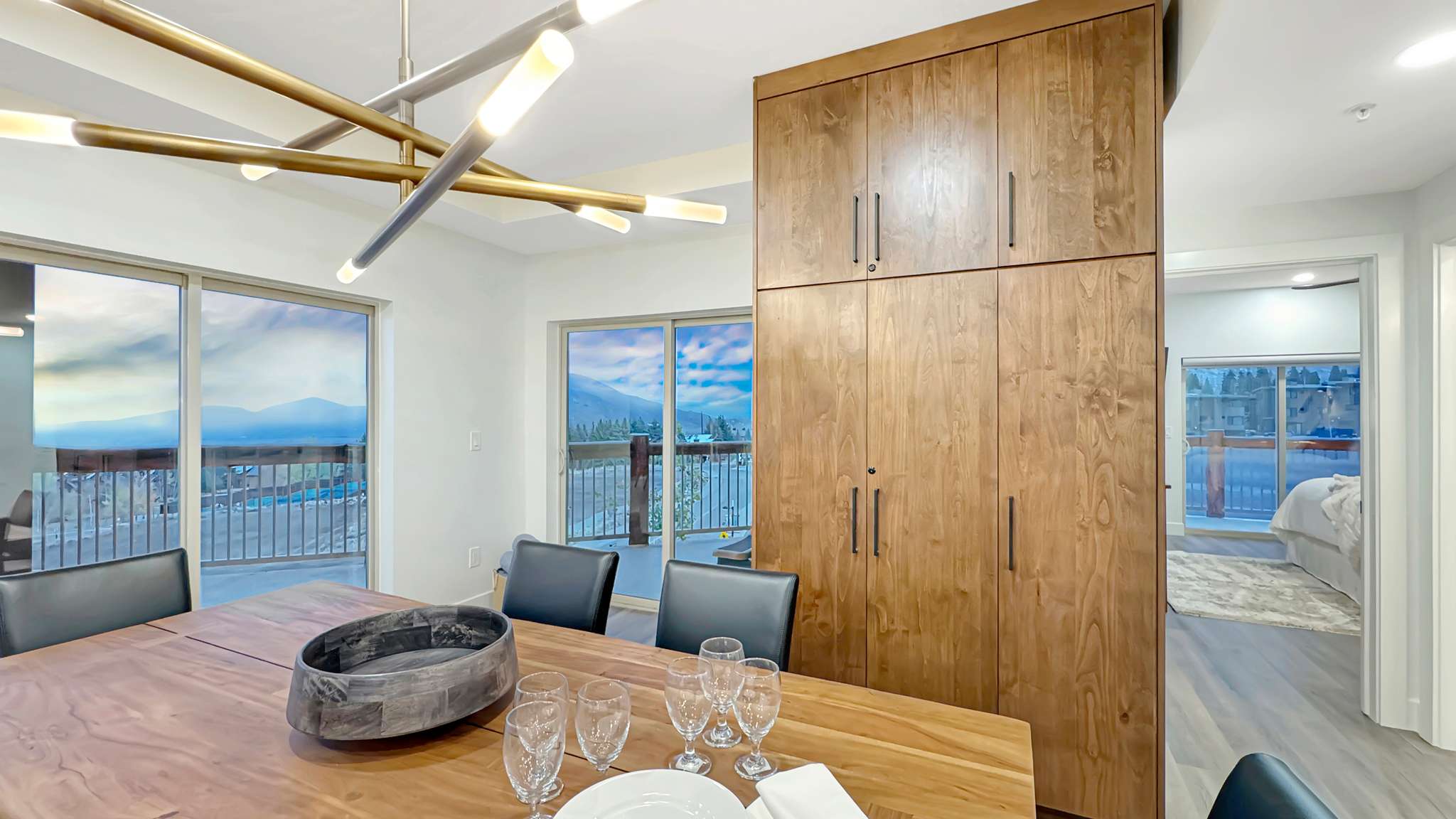 Dining area featuring a modern linear chandelier with brass accents, large sliding glass doors opening to a balcony, a full-height wood-finish storage unit with minimalist hardware, and light wood-finish flooring