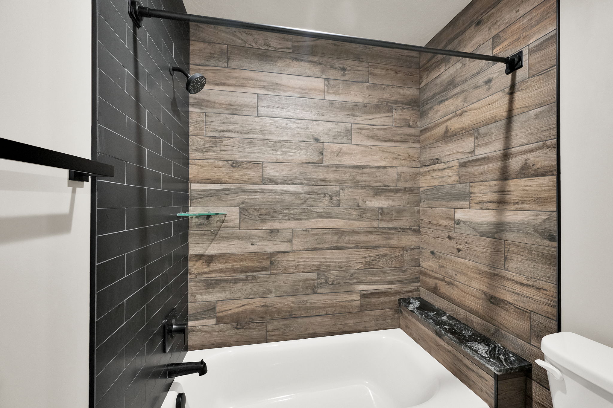 Bathtub shower featuring wood-look tile surround, dark subway tile accent, a built-in bench with a dark countertop, and a rainfall showerhead