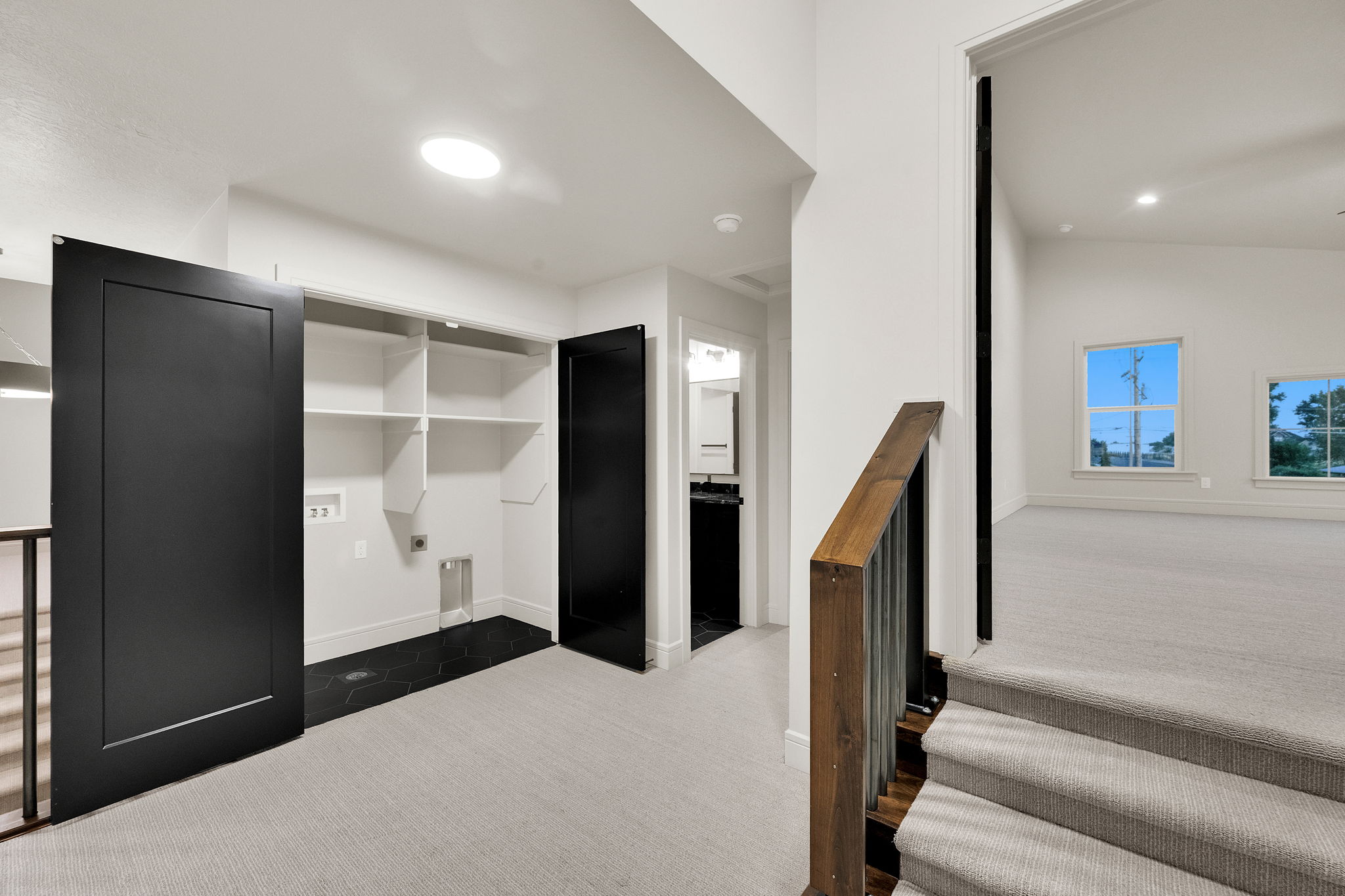Upper-level landing featuring a laundry area with black hexagon floor tiles and built-in shelving, a wood and metal stair railing, and a carpeted room with multiple windows
