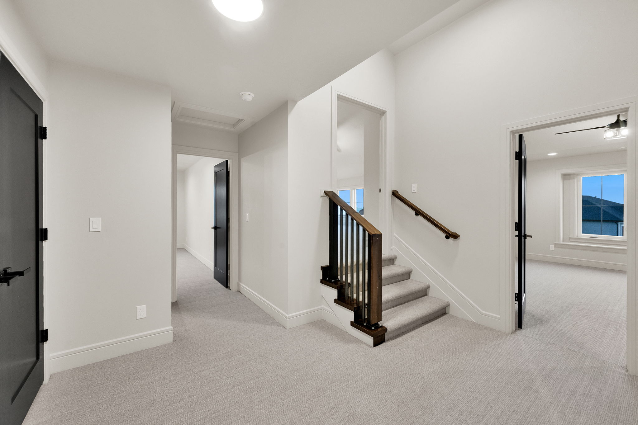 Upper-level hallway featuring a carpeted staircase with wood railing and dark balusters, light gray carpeting, crisp white walls, and dark-paneled doors with black hardware
