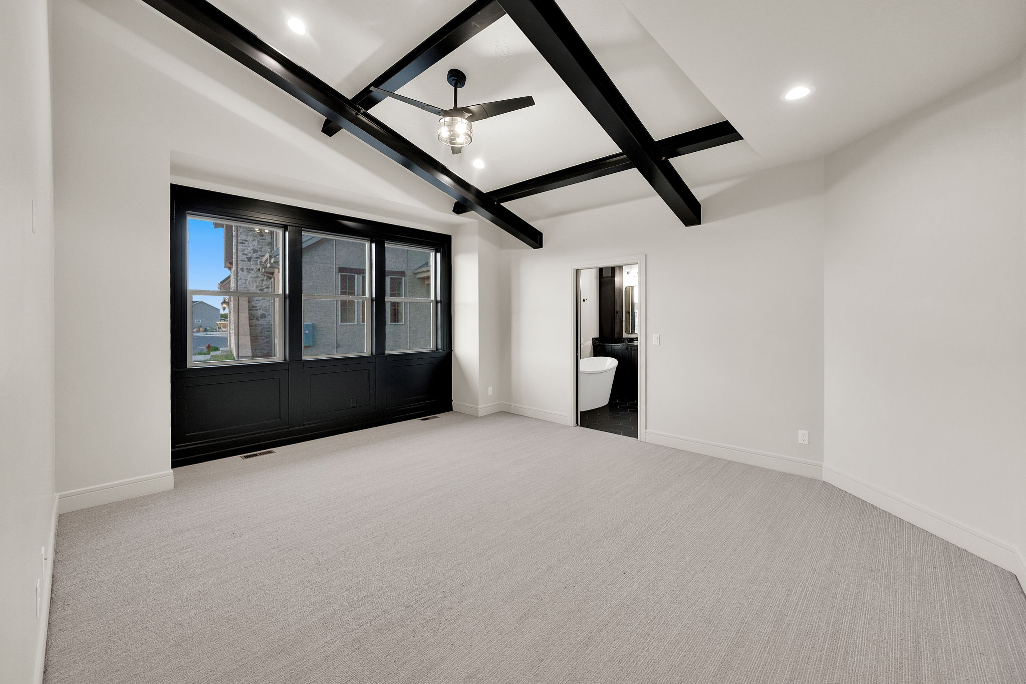 Spacious room featuring a vaulted ceiling with exposed black beams, light-toned carpeting, and a large multi-pane window with black framing