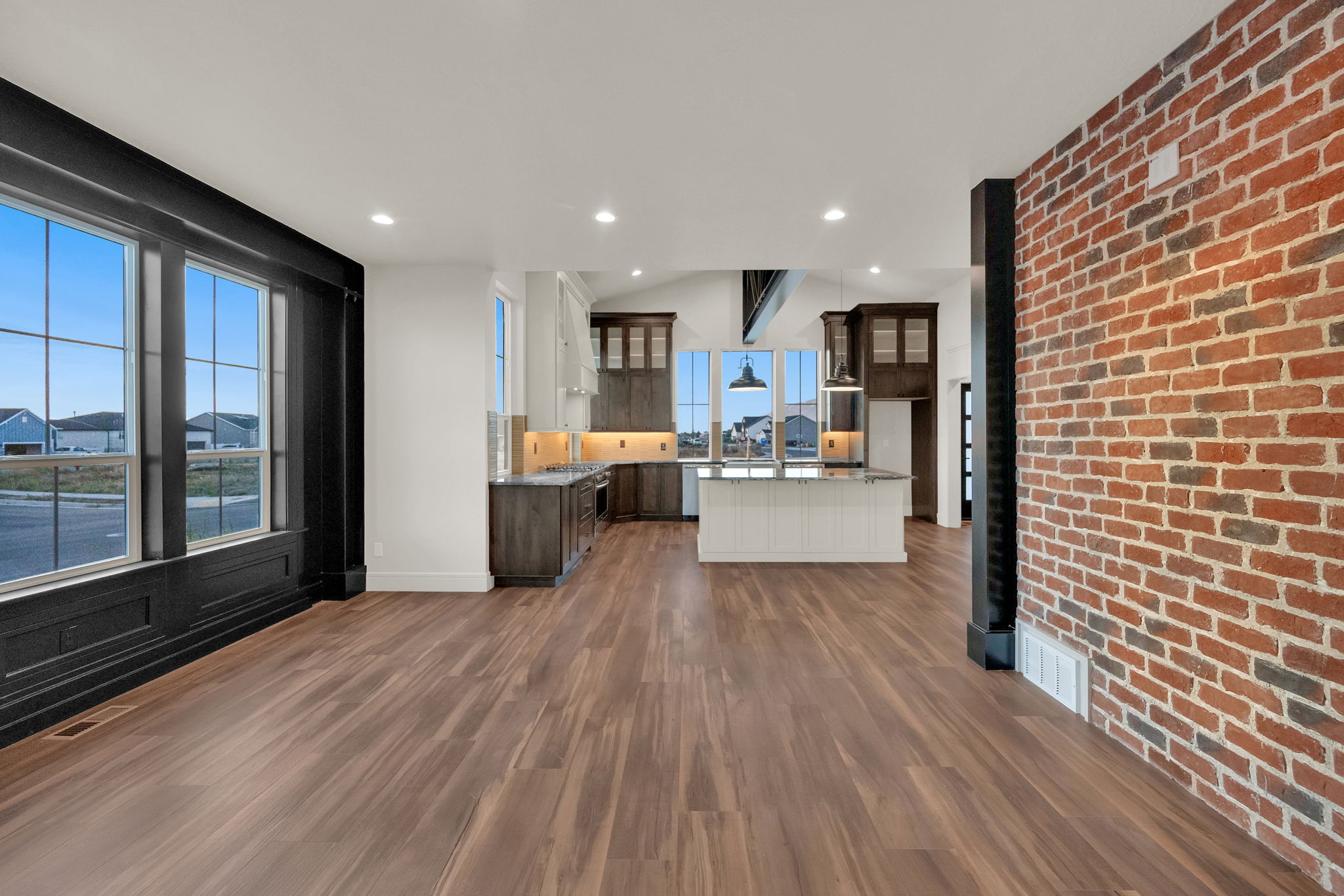 Spacious open-concept interior featuring wood-finish flooring, a prominent exposed brick wall, and extensive windows with dark trim