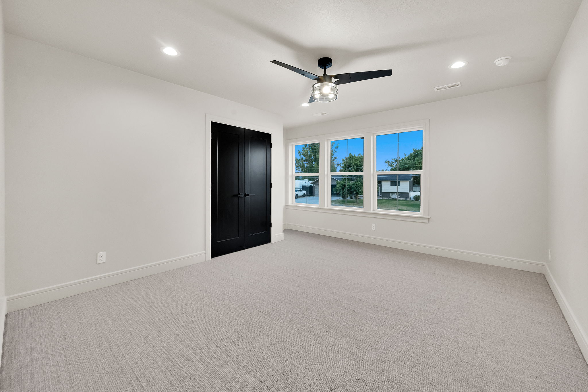 Spacious room featuring light-toned carpeting, ample natural light from three large windows, a contemporary ceiling fan with integrated lighting, recessed ceiling lights, and double panel doors