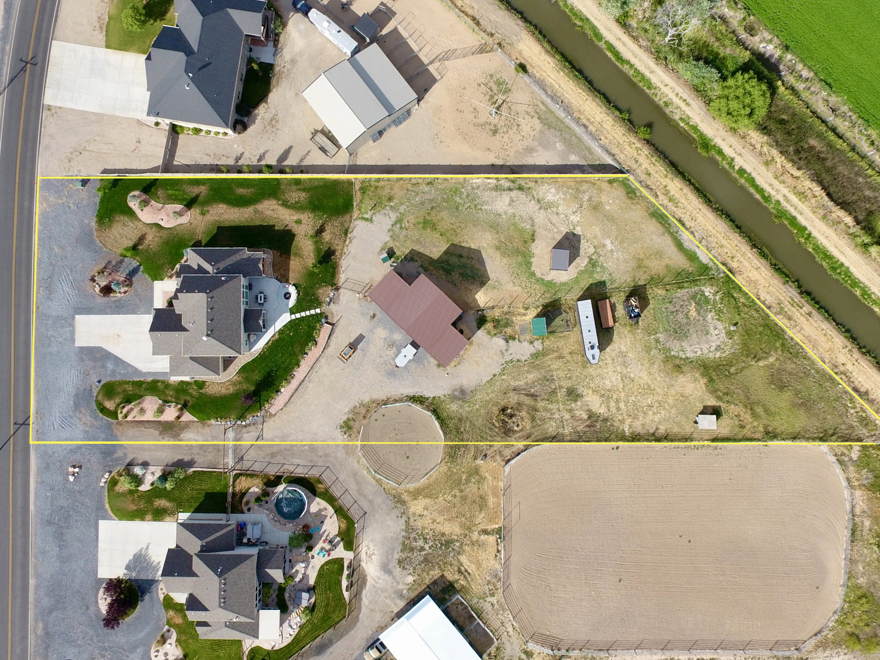 Aerial view showcasing a rectangular property featuring a primary residence with a concrete driveway, an additional structure with a brown metal roof, and an expansive sand arena