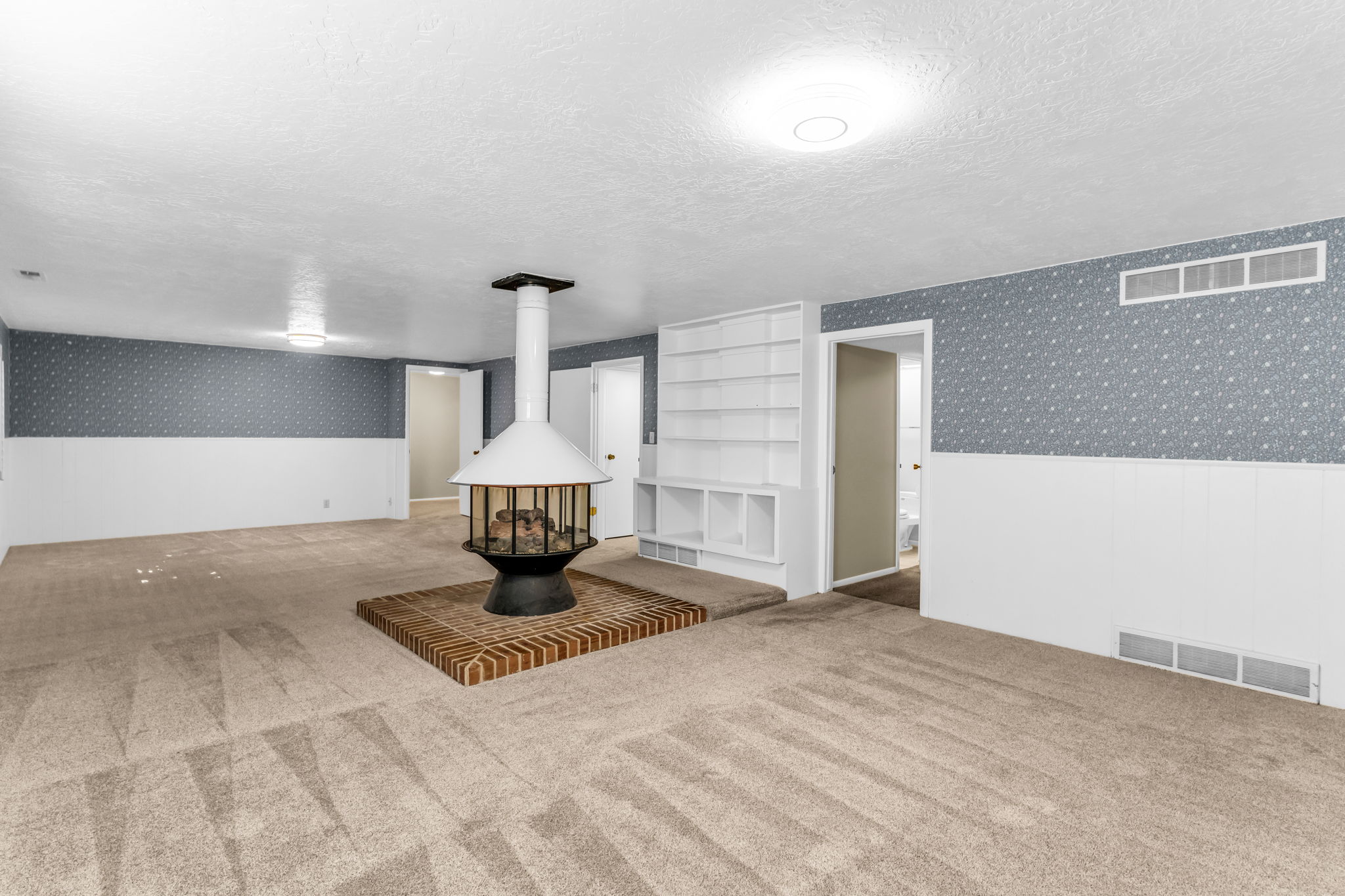 Freestanding fireplace with a brick hearth, wall-to-wall carpeting, built-in shelving, white paneled wainscoting, and patterned wall covering