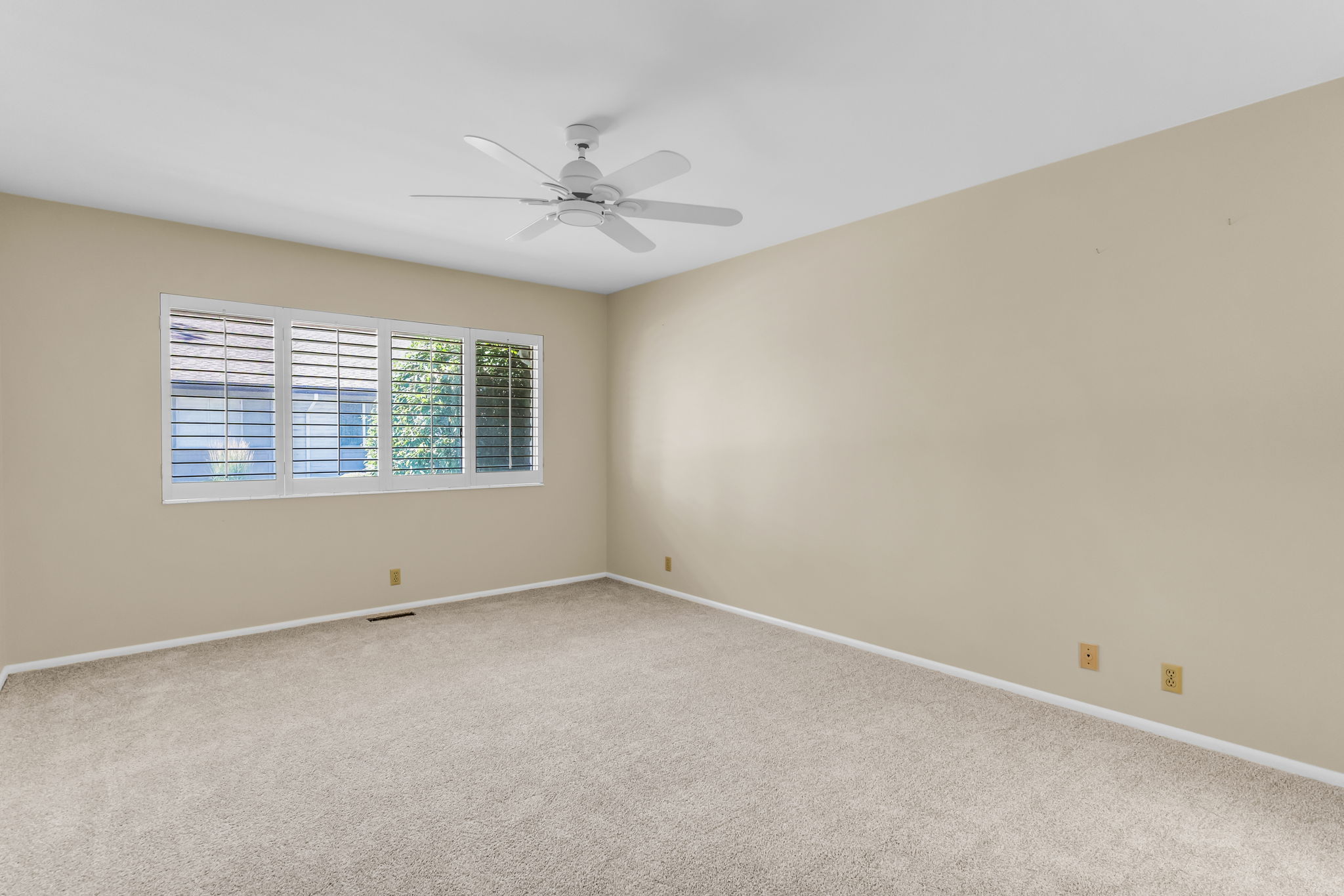 Carpeted room featuring a ceiling fan, a window with plantation shutters, light beige wall paint, and white baseboards
