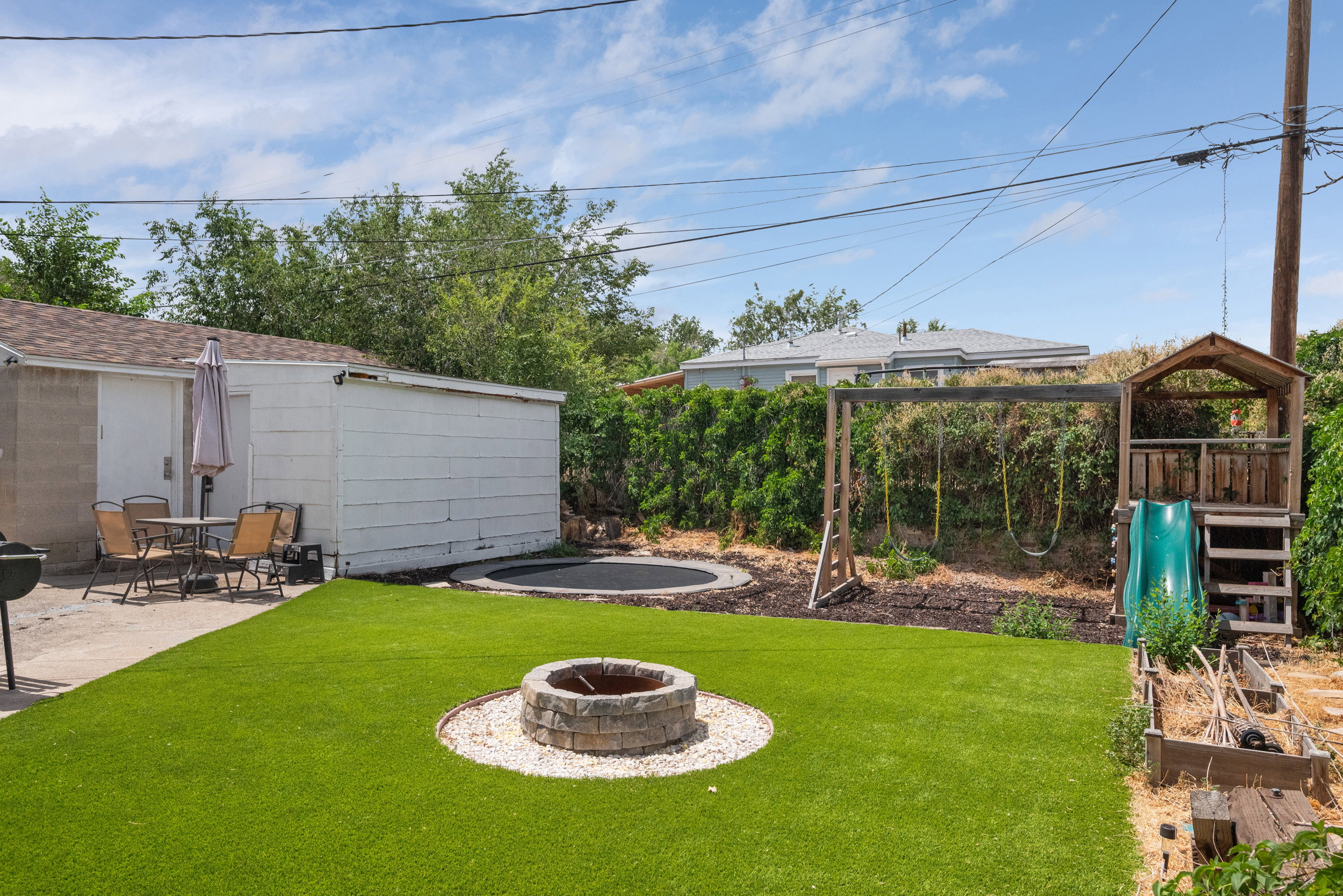 Landscaped backyard featuring an artificial turf lawn, a stone fire pit with white gravel surround, and a concrete patio