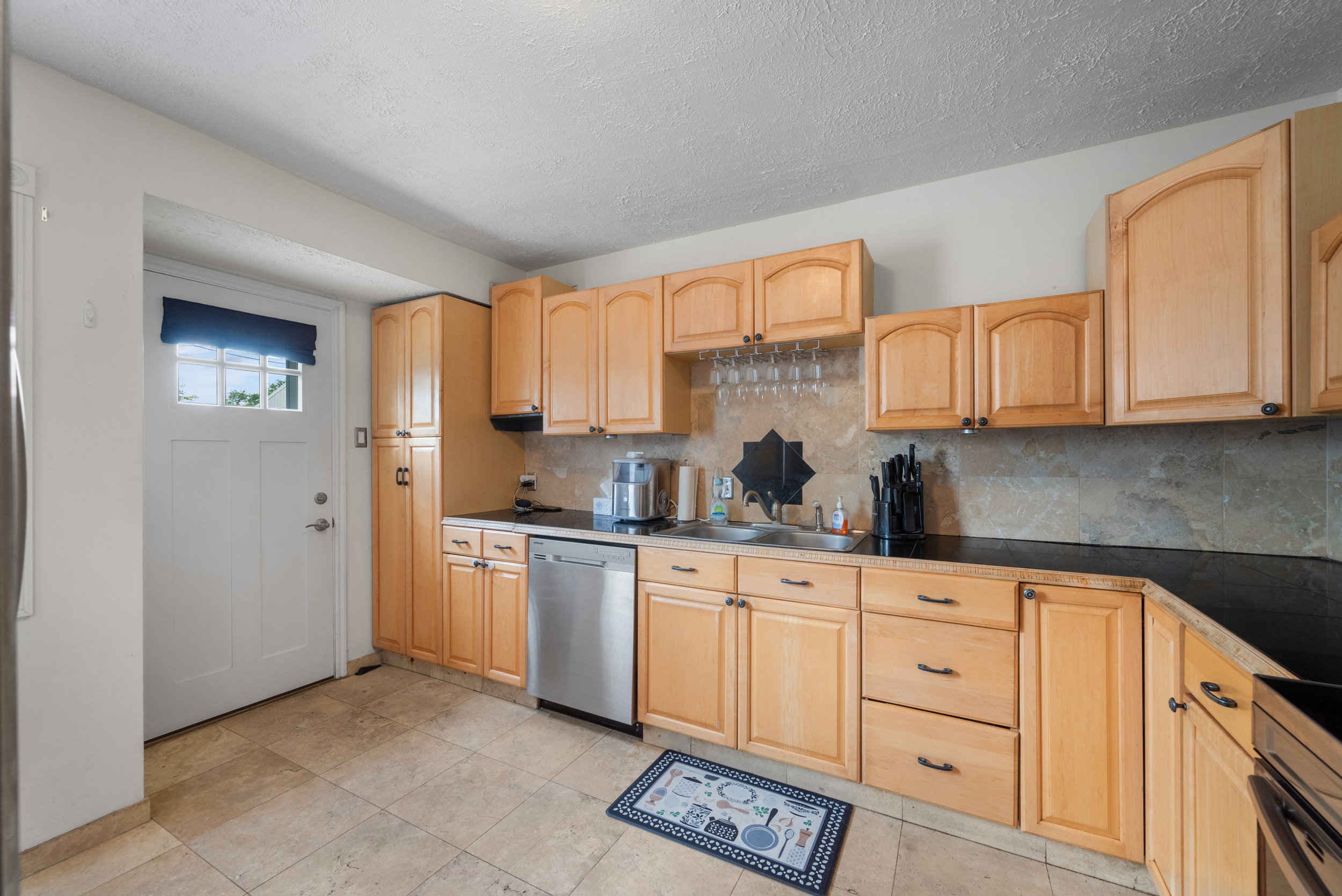 Kitchen featuring light wood cabinetry, dark countertops, a stainless steel dishwasher, an undermount sink, and stone-finish tile flooring