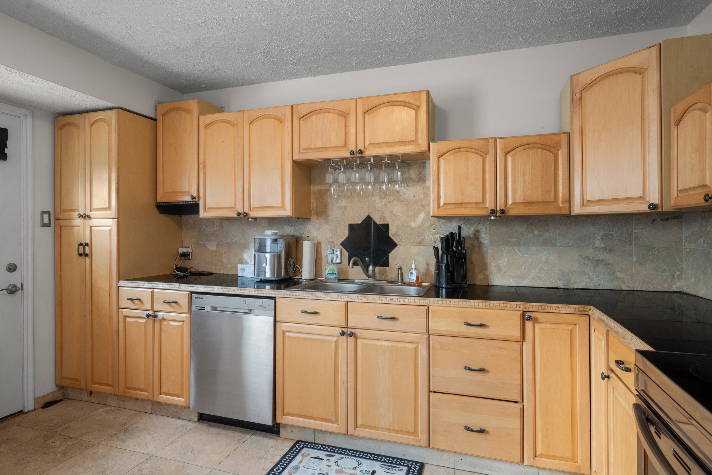 Kitchen featuring light wood cabinetry with arched top panels, a stainless steel dishwasher, black countertops, and a stone-finish backsplash