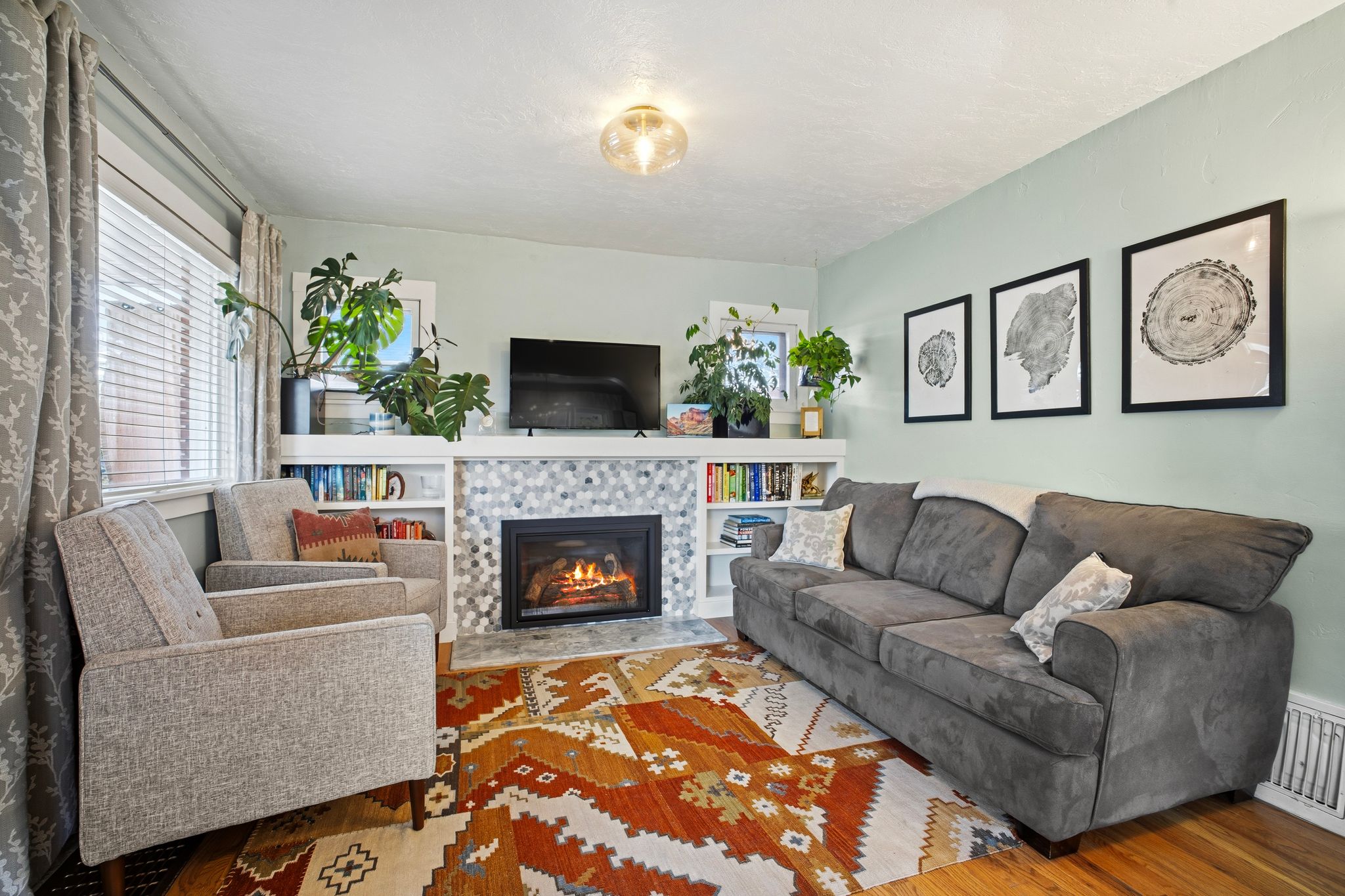 Living space featuring hardwood flooring, a fireplace with a hexagonal tile surround, built-in shelving, and a window with blinds and drapery