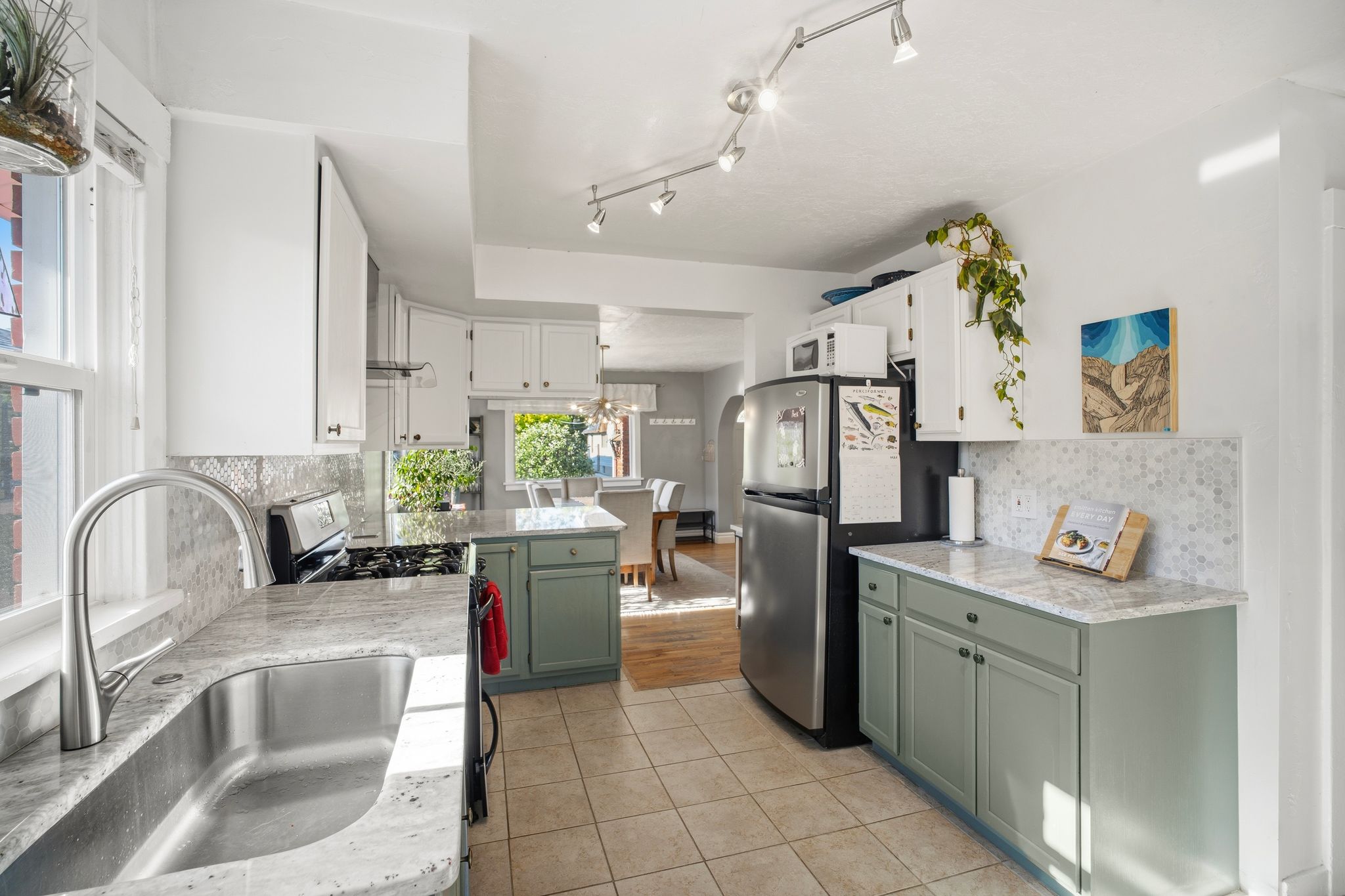 Bright kitchen featuring a stainless steel sink with a gooseneck faucet, light-toned countertops, a patterned tile backsplash, and two-tone cabinetry with upper white and lower sage green finishes