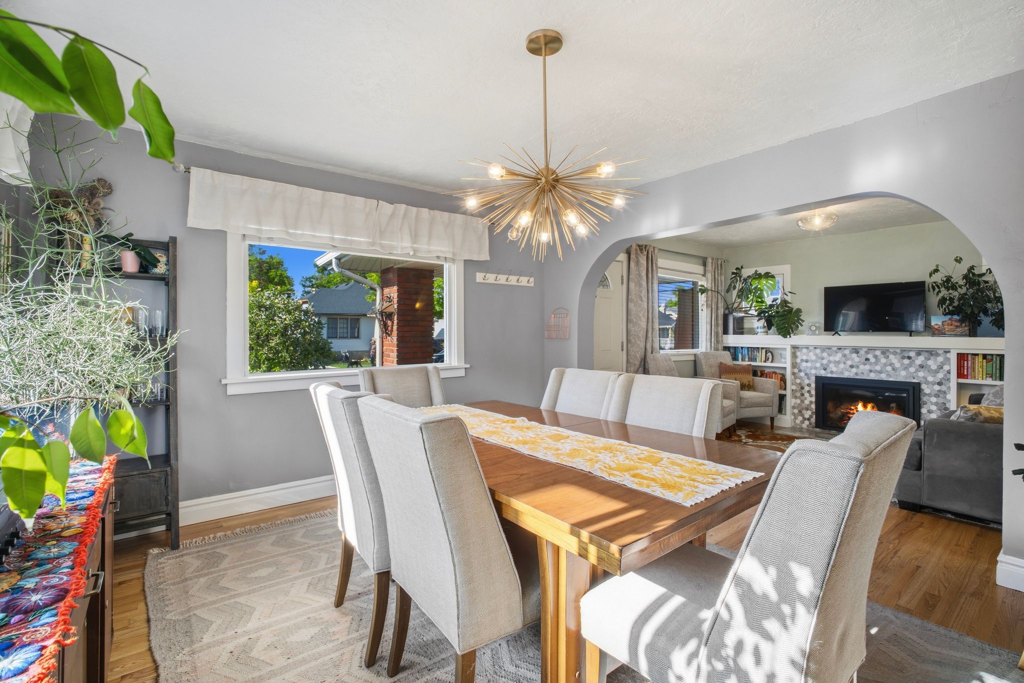 Dining area featuring hardwood flooring, a modern sputnik chandelier, and an arched doorway leading to a living space with a tiled fireplace and built-in shelving