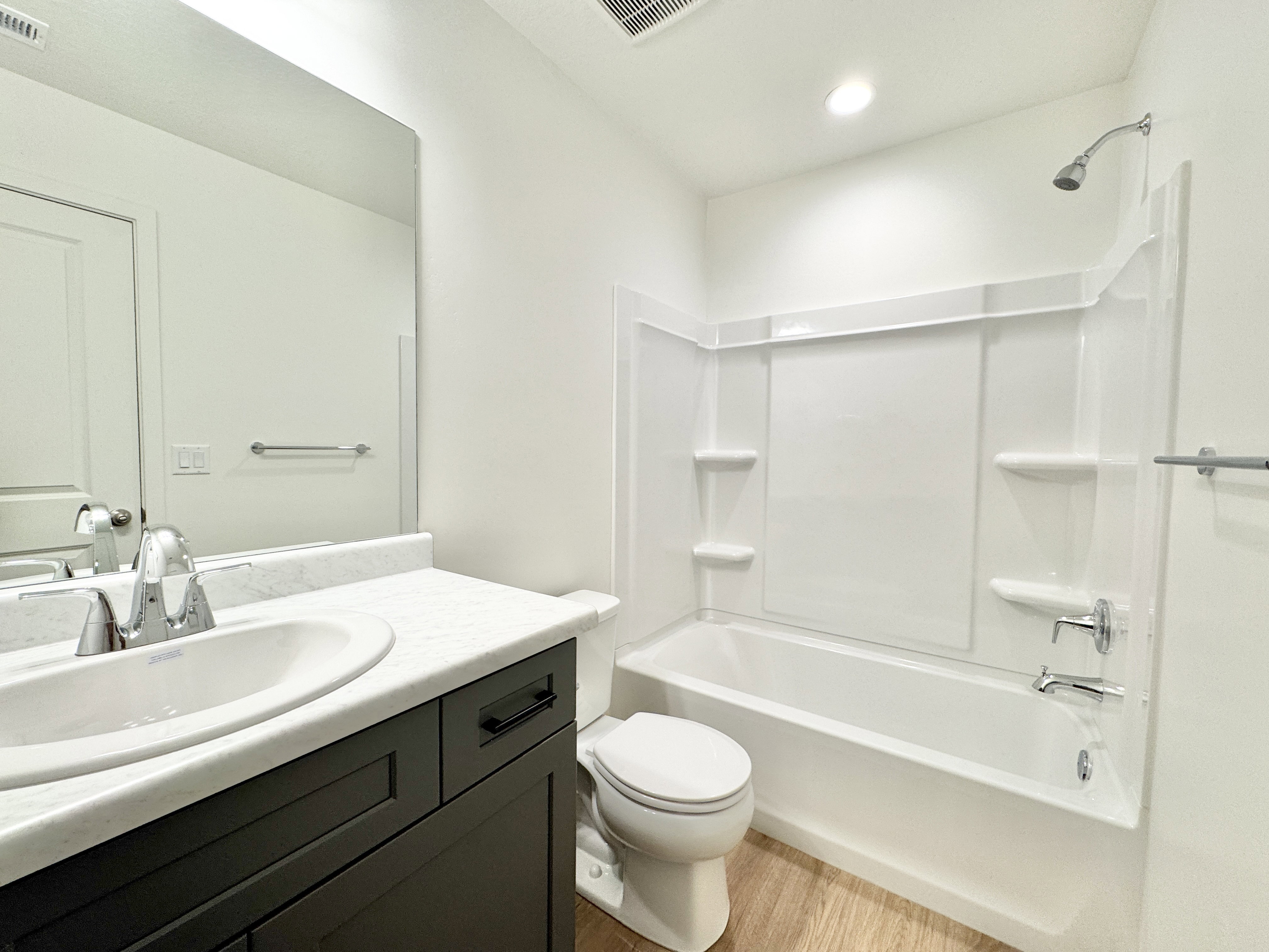 Bathroom featuring a white tub and shower combination with integrated shelving, a dark wood-finish vanity with a white countertop and integrated sink, and wood-finish flooring