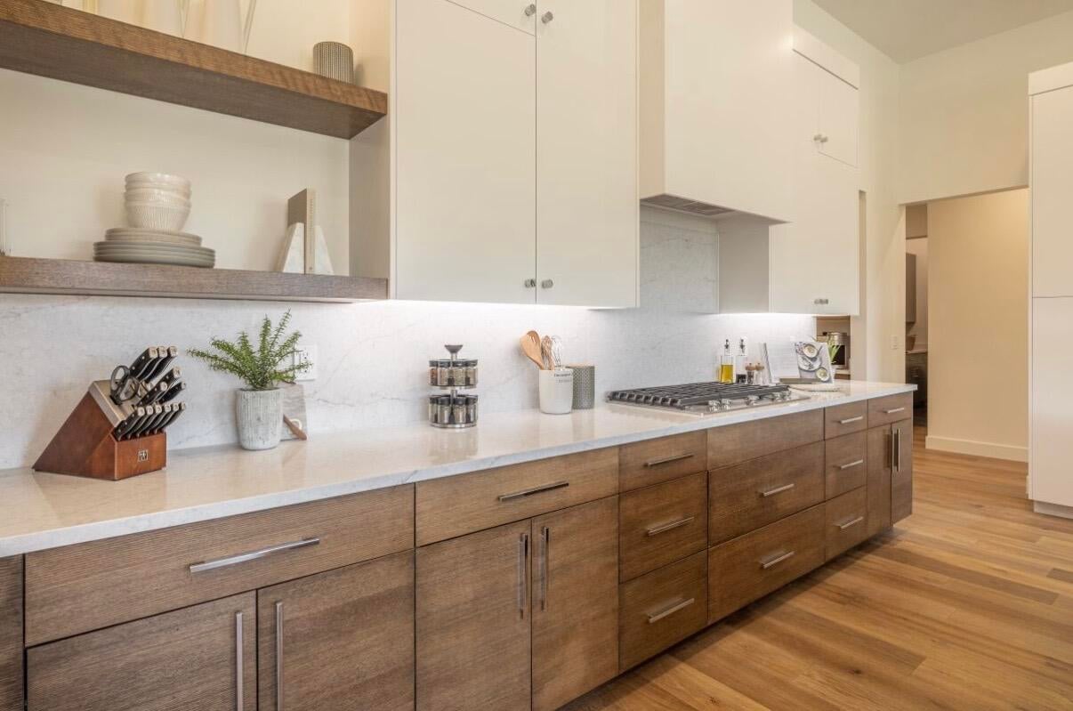 Modern kitchen featuring a gas range, white countertops, full-height backsplash, wood-finish flooring, and two-tone cabinetry with upper white cabinets and lower wood-finish cabinets