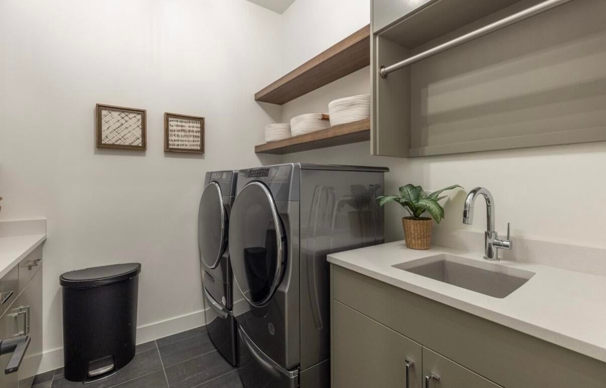 Laundry room featuring a built-in utility sink with a chrome faucet, light-toned countertop, wood-finish floating shelves, and grey cabinetry with polished hardware