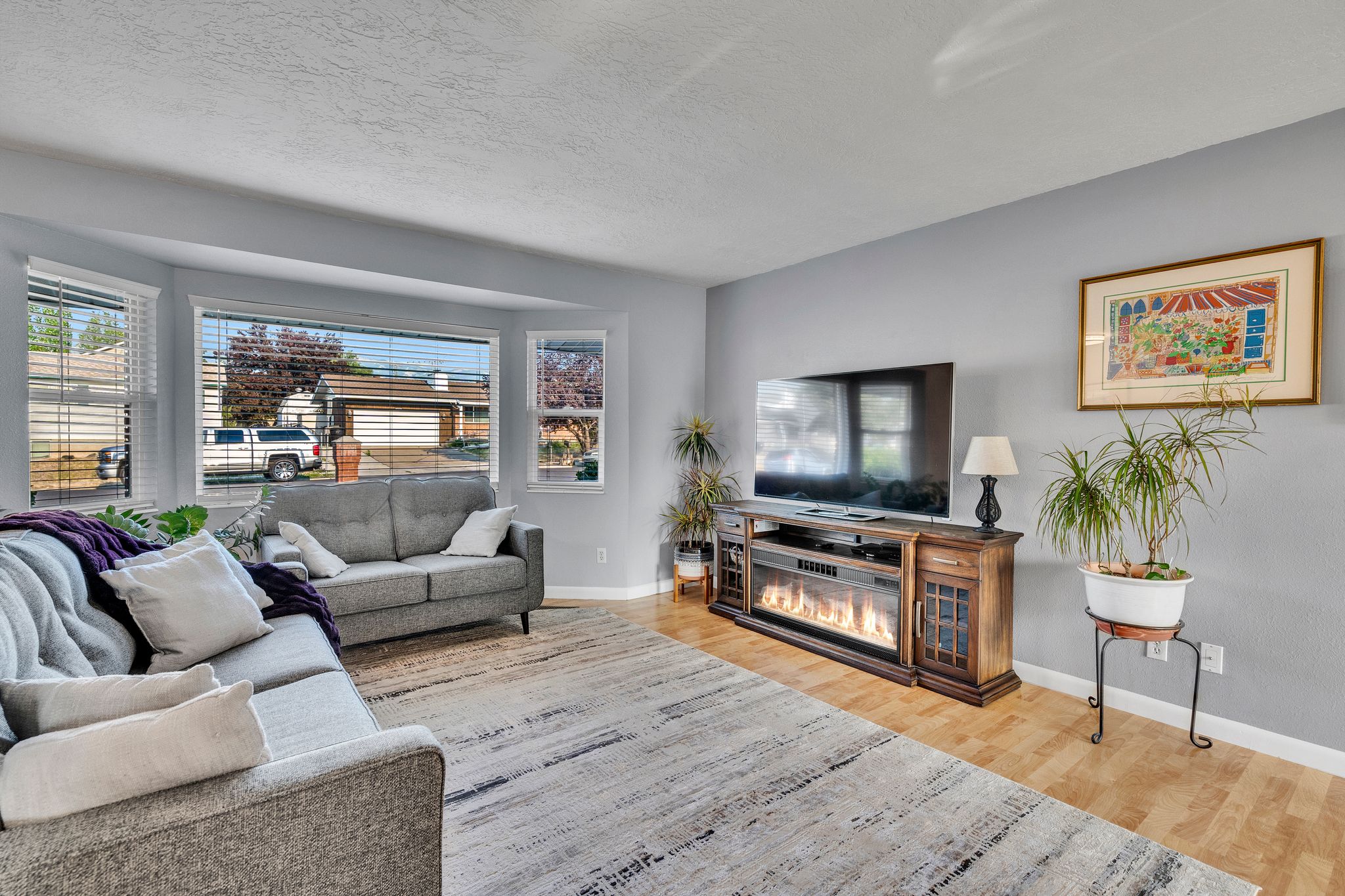 Living area featuring light wood-finish flooring, a contemporary electric fireplace media console, and a large bay window with horizontal blinds