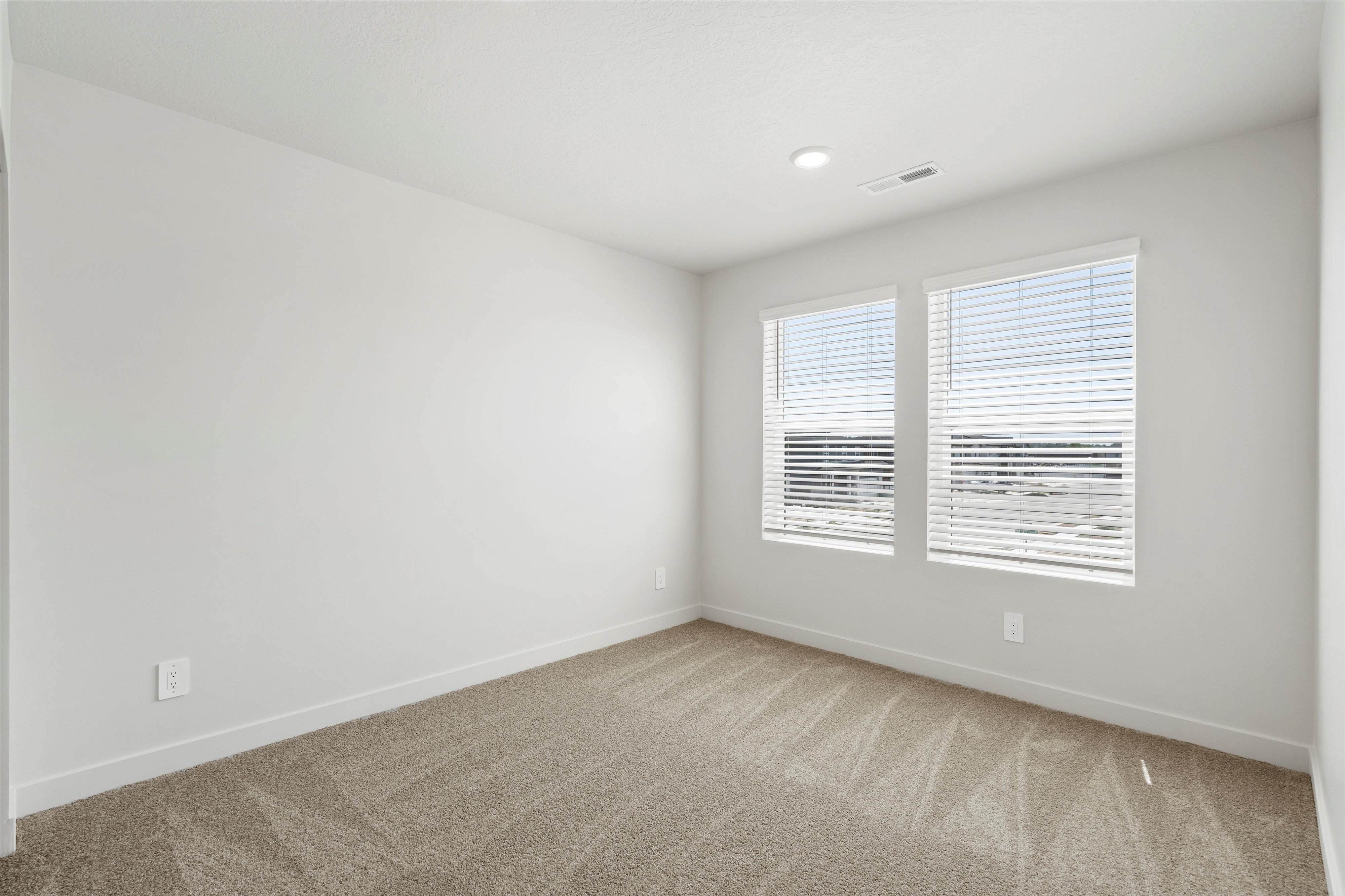 **DISCLAIMER** Interior package may vary. Neutral-toned room featuring a light-colored carpet, white baseboards, and two windows with blinds