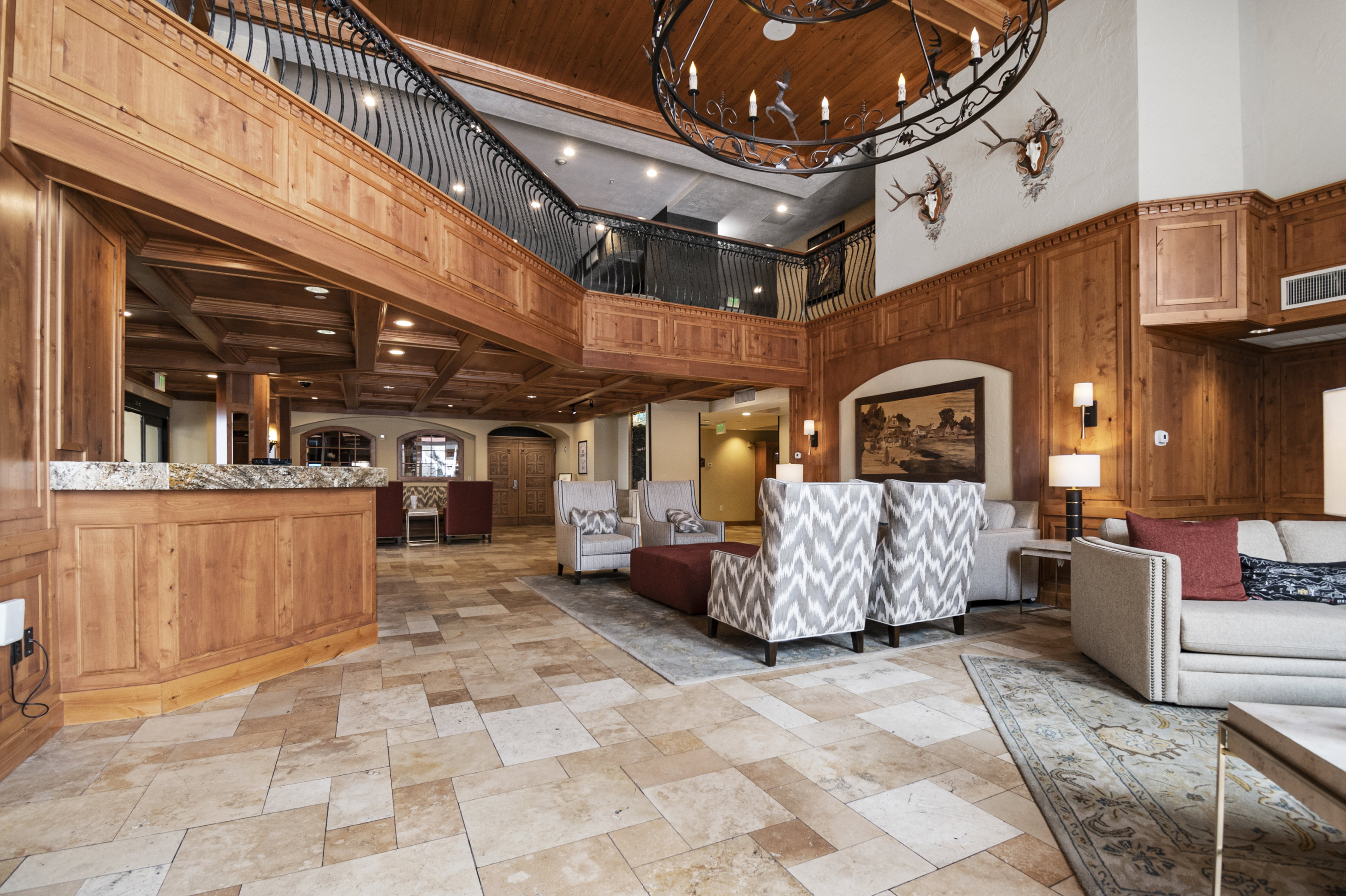 Expansive foyer featuring a grand staircase with wrought iron railing, intricate wood paneling, a large decorative chandelier, natural stone flooring, and a reception counter with a stone surface