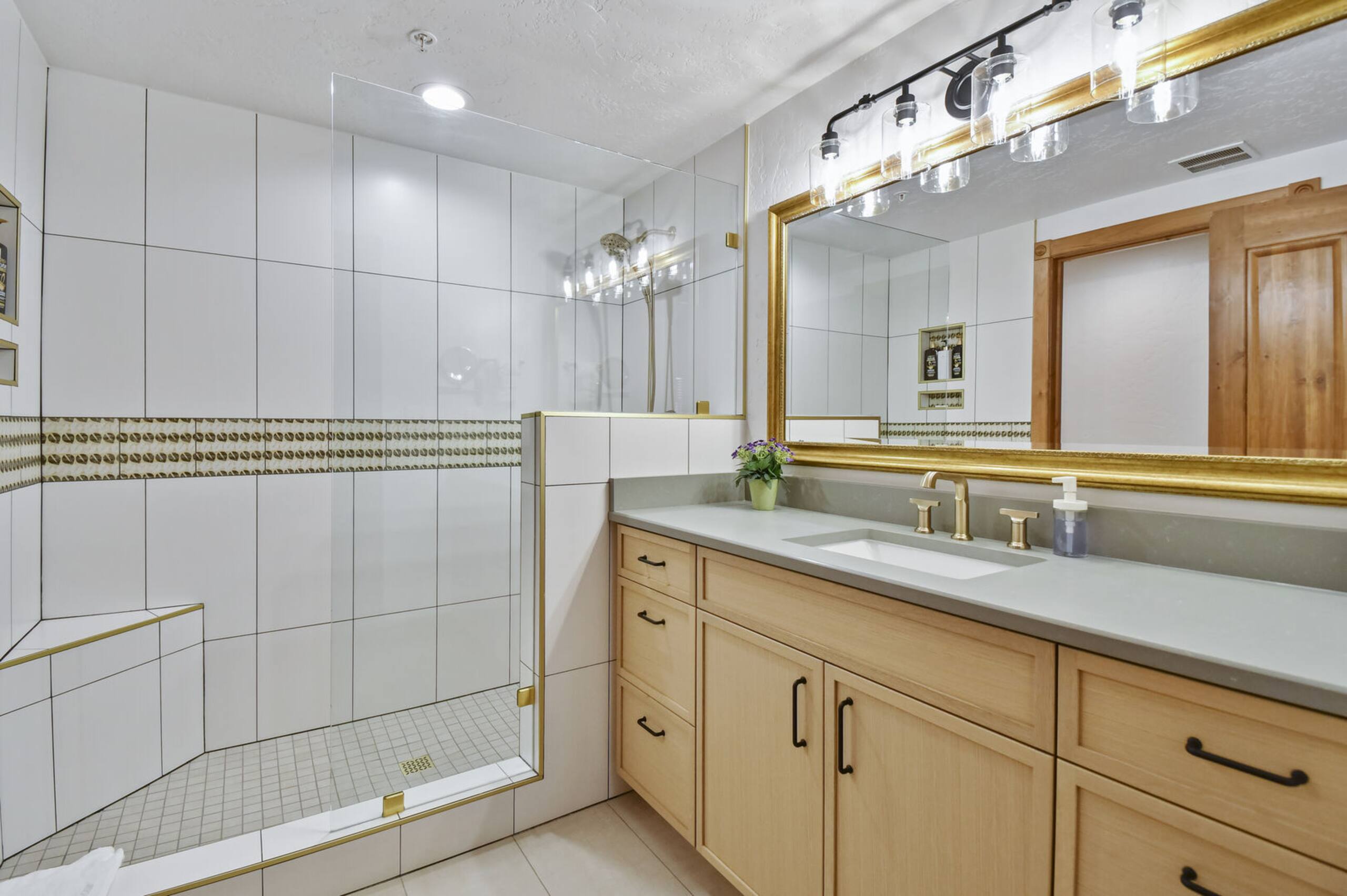 Spacious bathroom featuring a frameless glass shower enclosure with a built-in bench, light wood-finish vanity with ample storage, a large gold-framed mirror, a single undermount sink, and a multi-bulb vanity light fixture