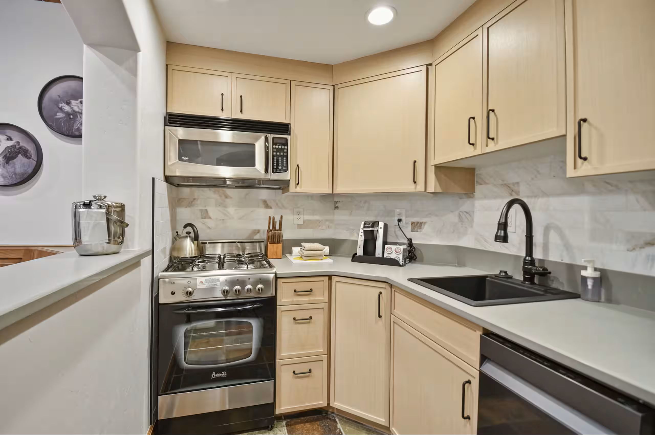 Compact kitchen featuring light wood-finish cabinetry, stainless steel appliances, a black sink with a matte black faucet, and a stone-look backsplash