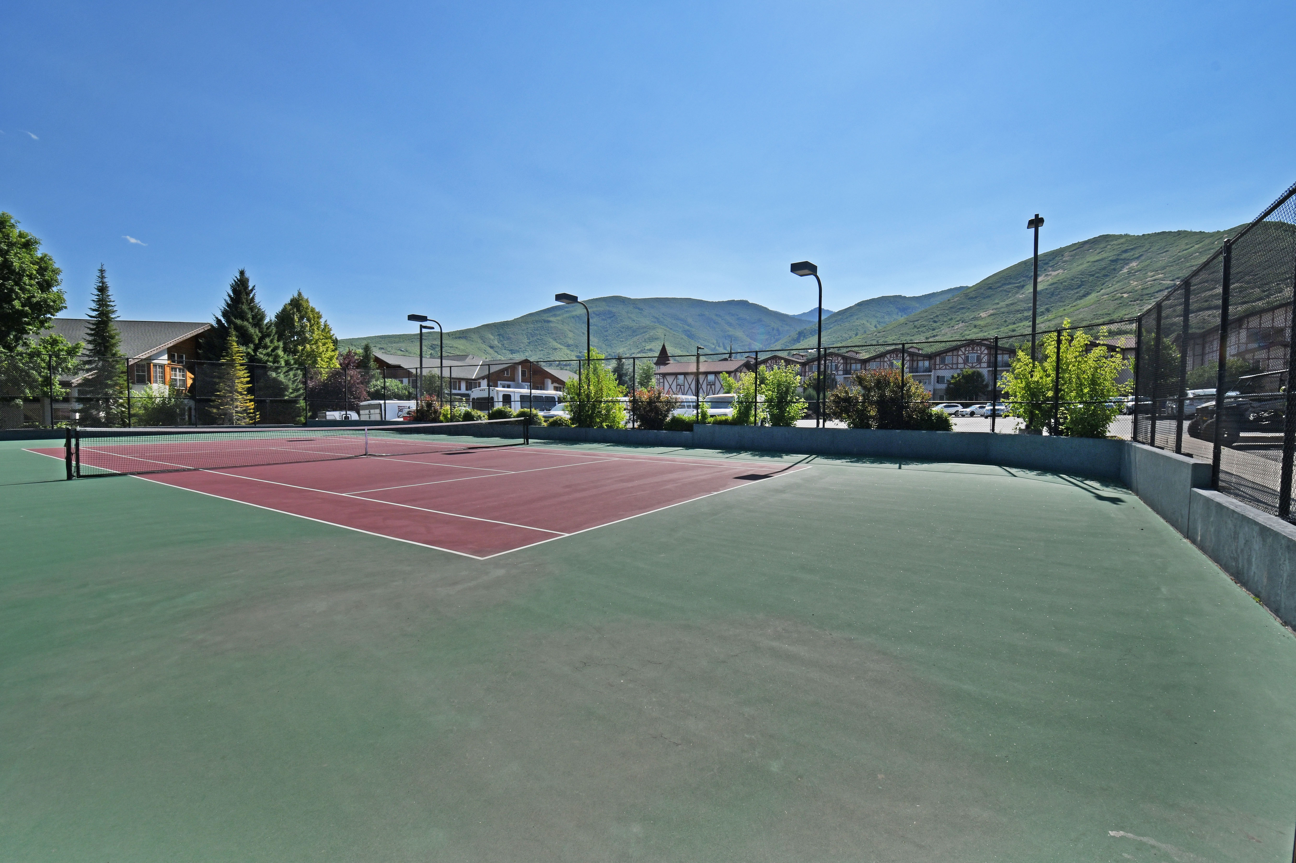 Hard surface tennis court featuring a green and red playing area, perimeter fencing, and overhead lighting