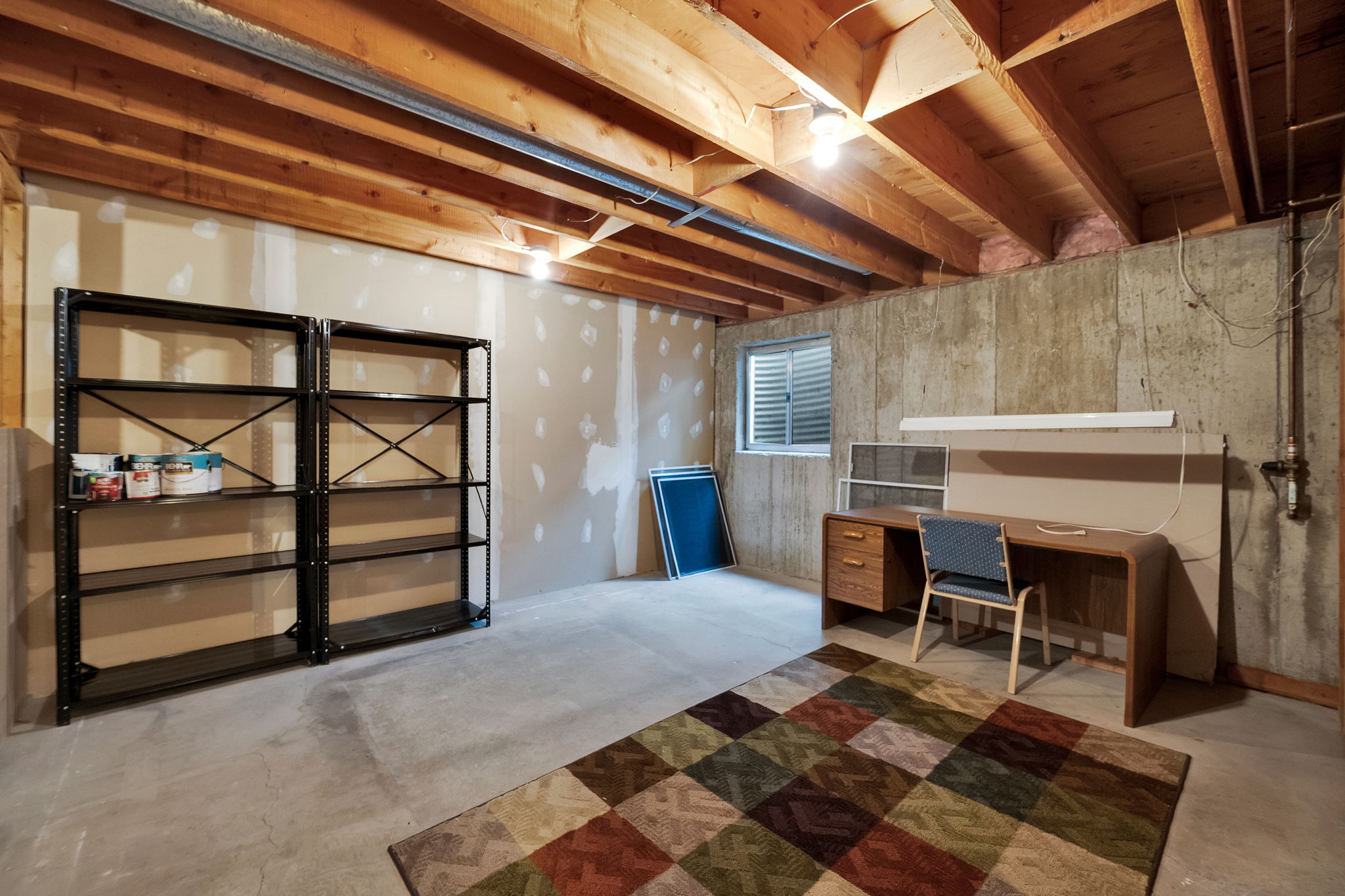 Unfinished basement area featuring exposed wooden ceiling joists, concrete flooring, and a window with a louvered well cover