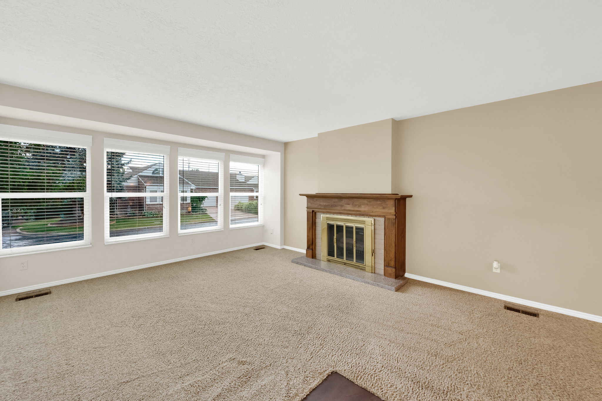 Living area featuring a classic wood mantel fireplace with a gold-toned insert, a gray stone hearth, beige carpeting, and multiple large windows with blinds