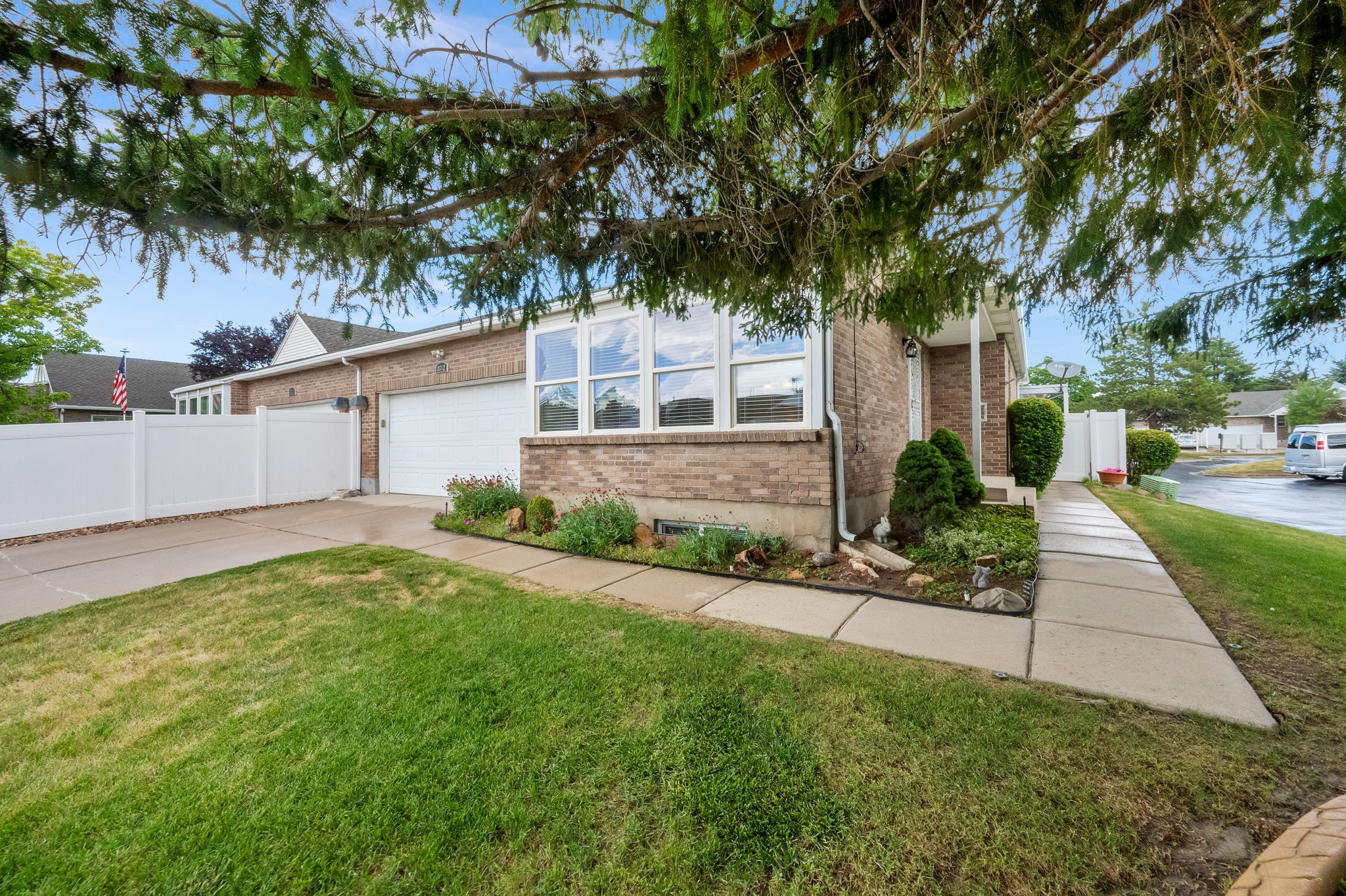 Brick facade residence with an attached garage, featuring a concrete driveway, a white vinyl fence, and a landscaped front yard with mature shrubbery