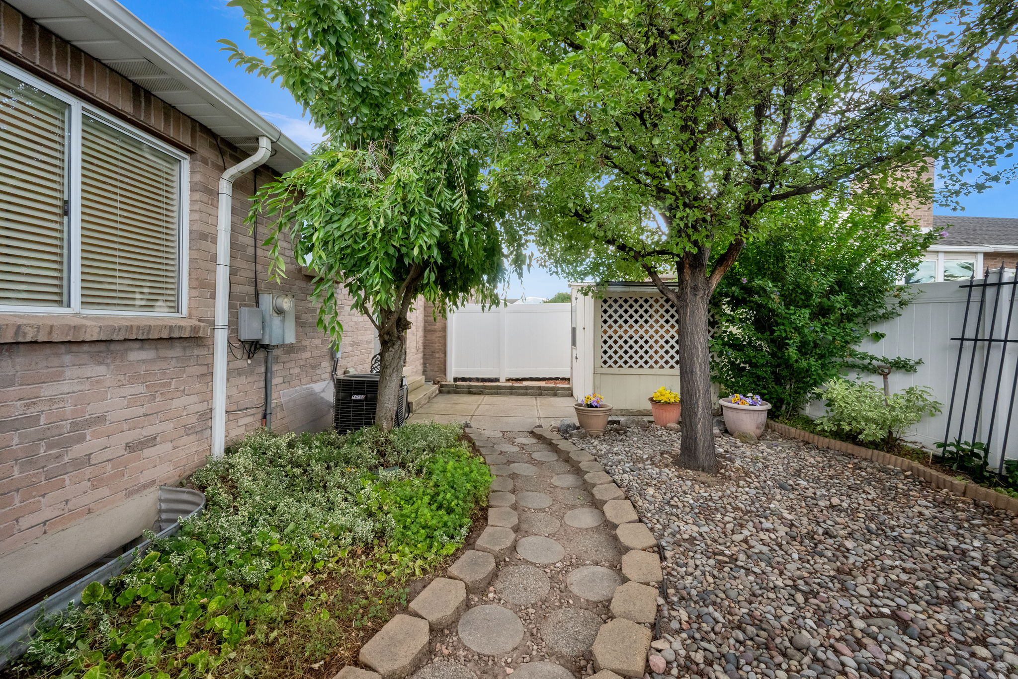 Brick exterior featuring a stone paver pathway, mature tree, established shrubbery, and a white privacy fence