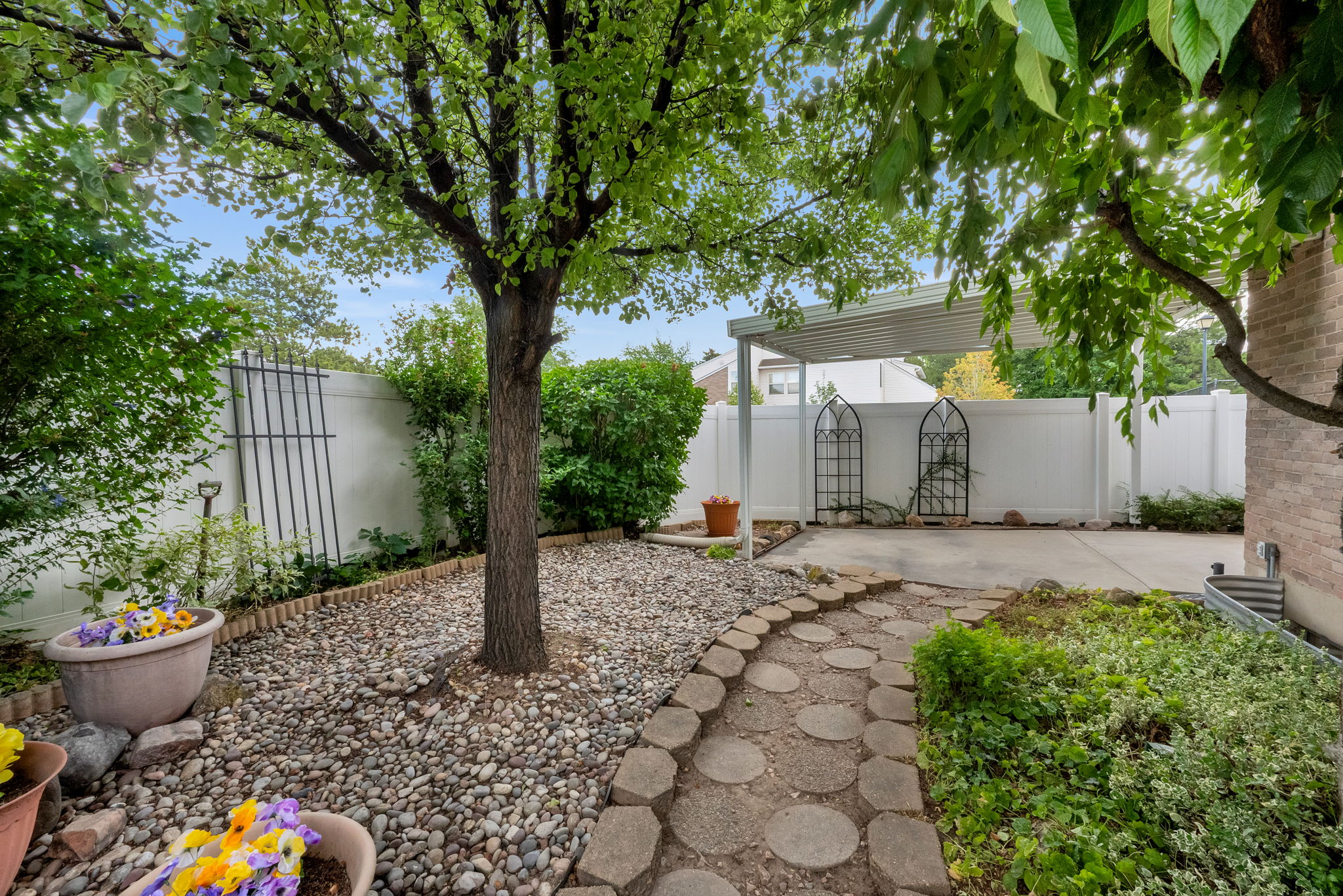 Landscaped backyard featuring a stone paver pathway, mature tree, gravel ground cover, and planted garden beds