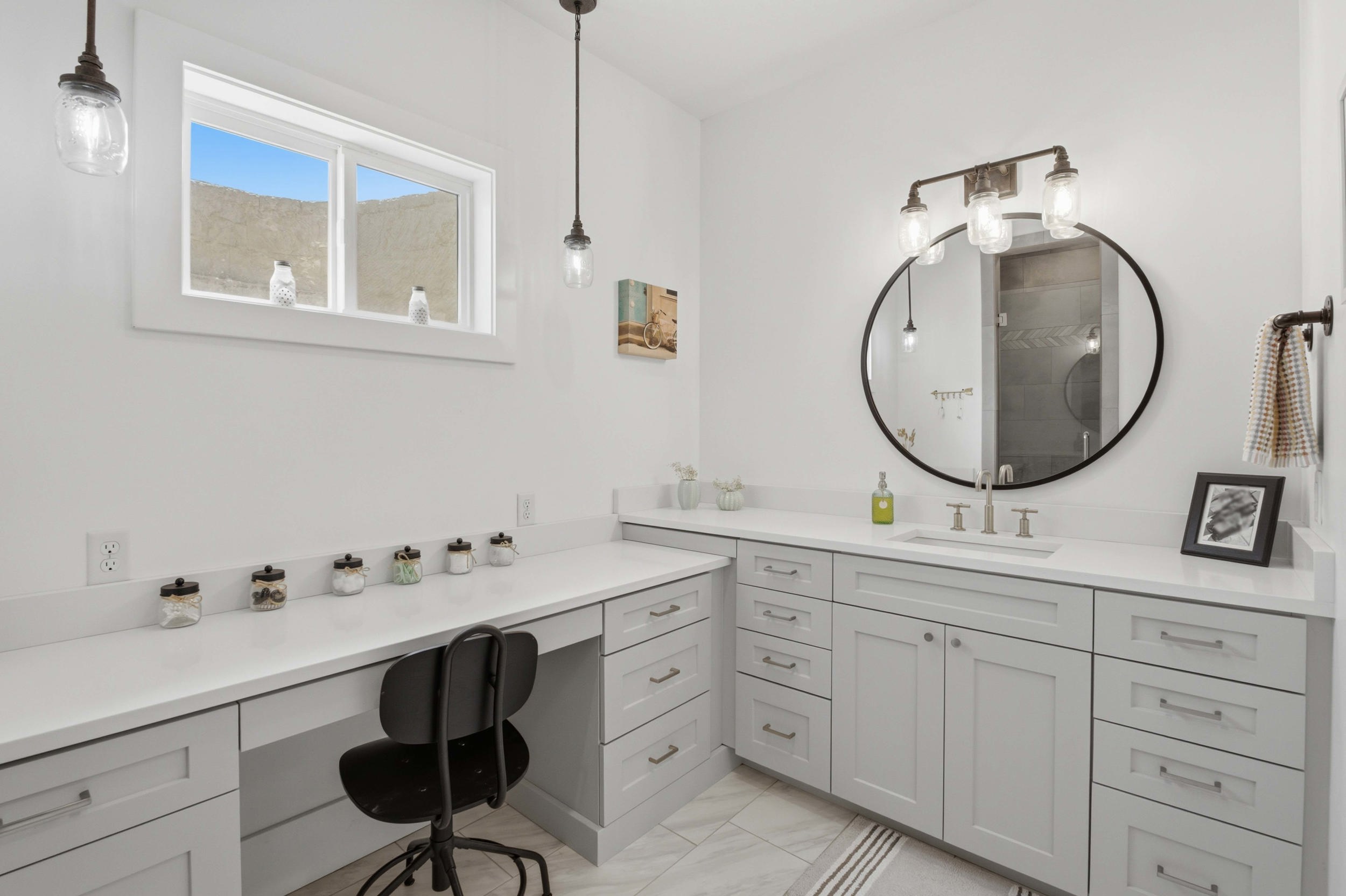 Spacious vanity area featuring light gray cabinetry, white countertops, built-in desk space, a round framed mirror, and contemporary sconce lighting