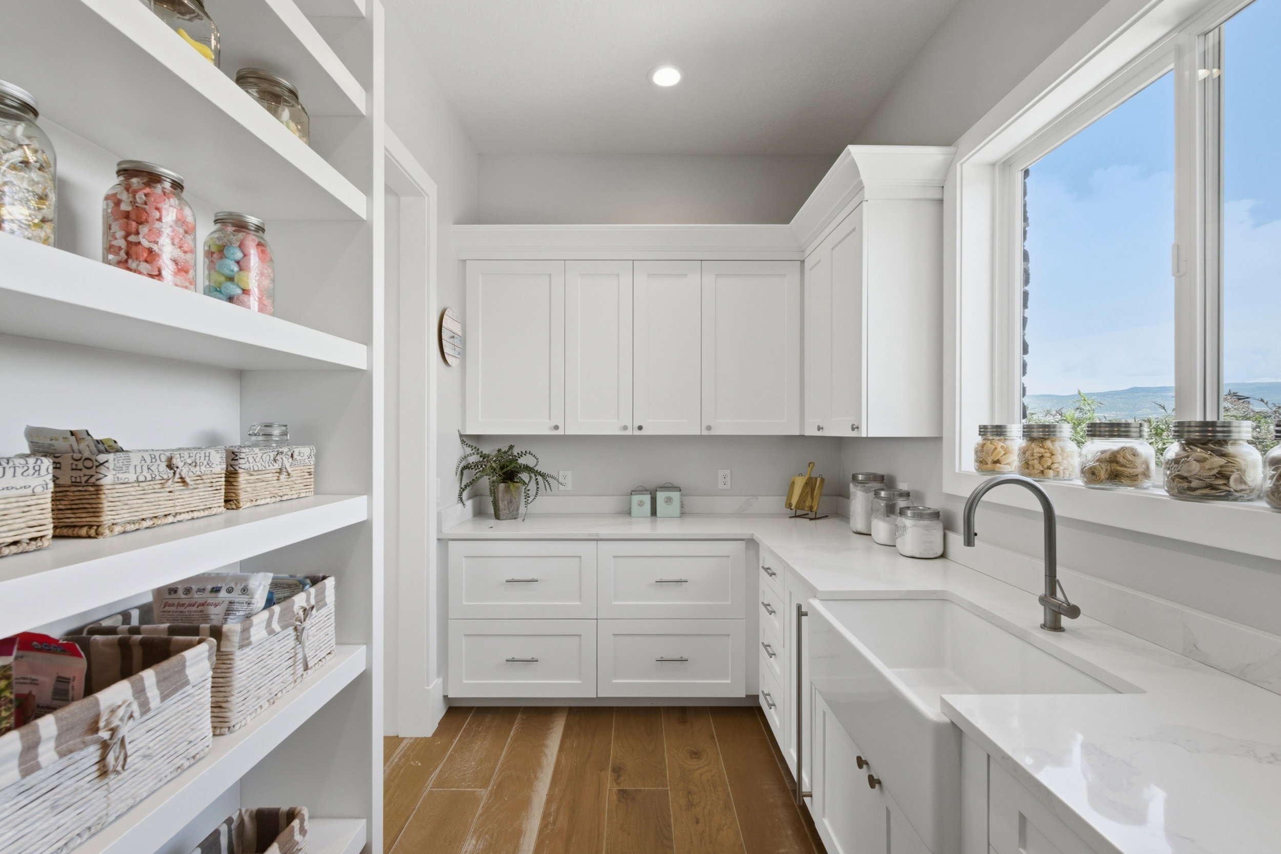 Well-appointed pantry featuring extensive built-in shelving, white shaker cabinetry, an integrated farmhouse sink with a gooseneck faucet, light-toned countertops, and wood-finish flooring