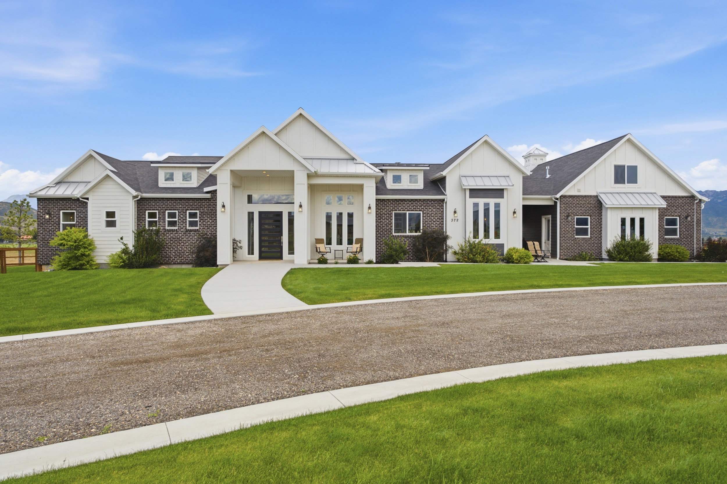 Contemporary farmhouse exterior featuring board and batten siding, dark brick accents, a gabled roofline, a prominent covered entry, and a gravel driveway with a concrete walkway