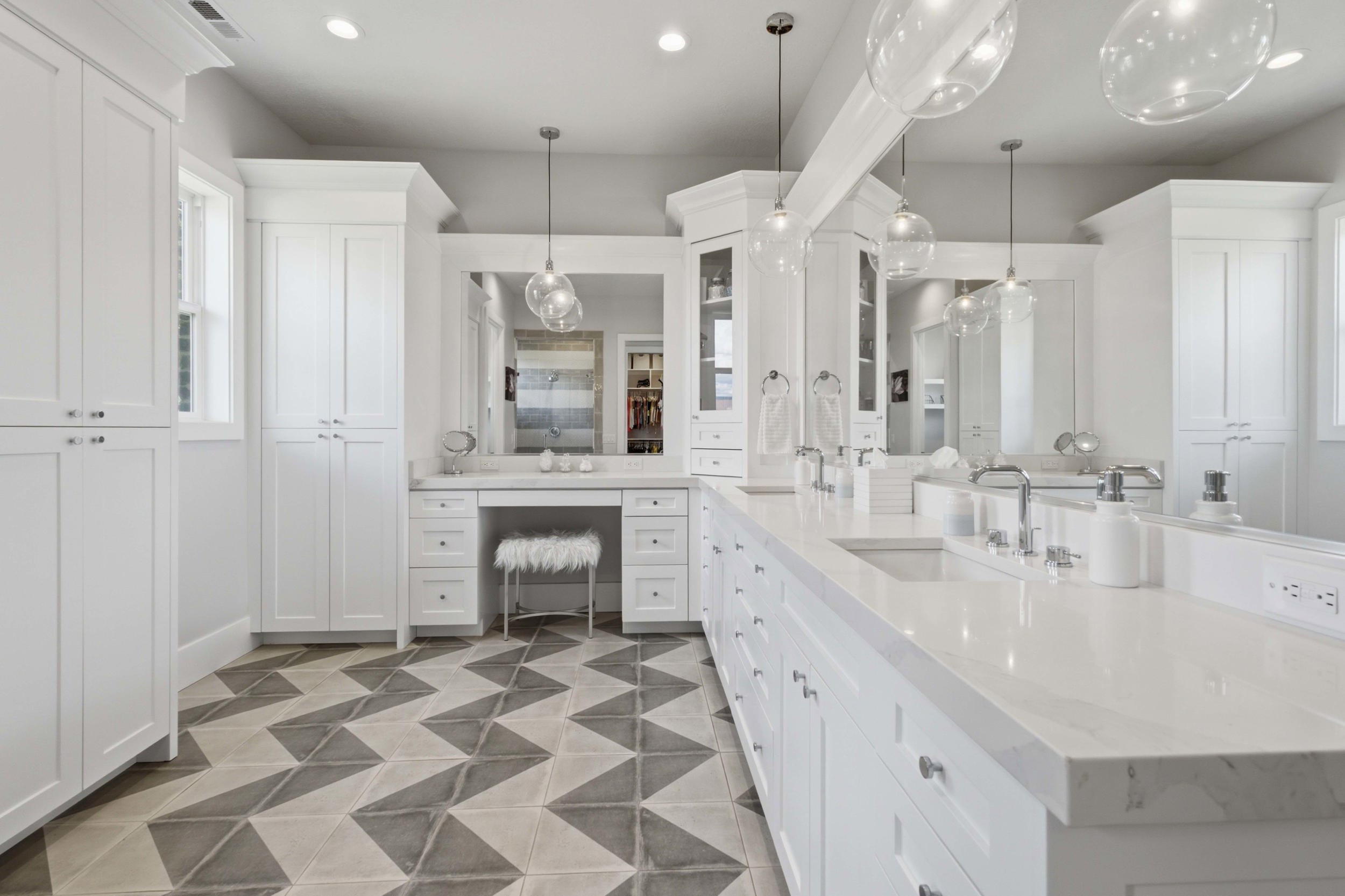 Spacious bathroom featuring an extended double vanity with undermount sinks and white countertops, custom white cabinetry for ample storage, a dedicated vanity area with mirror, contemporary pendant lighting, and geometric patterned floor tiling