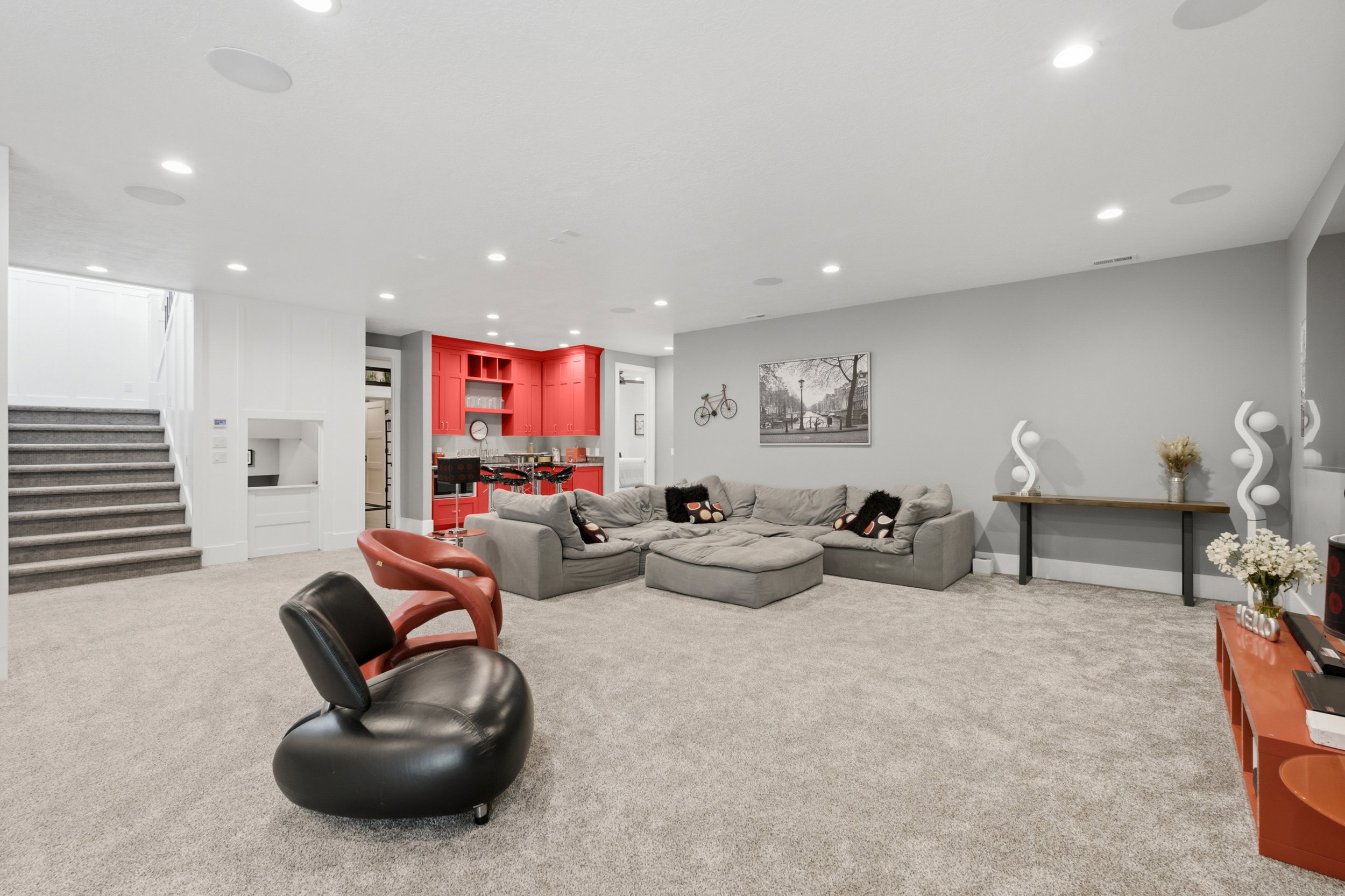 Spacious recreation room featuring light gray carpeting, recessed lighting, a built-in wet bar with red cabinetry, a built-in desk area, and a staircase with carpeted treads