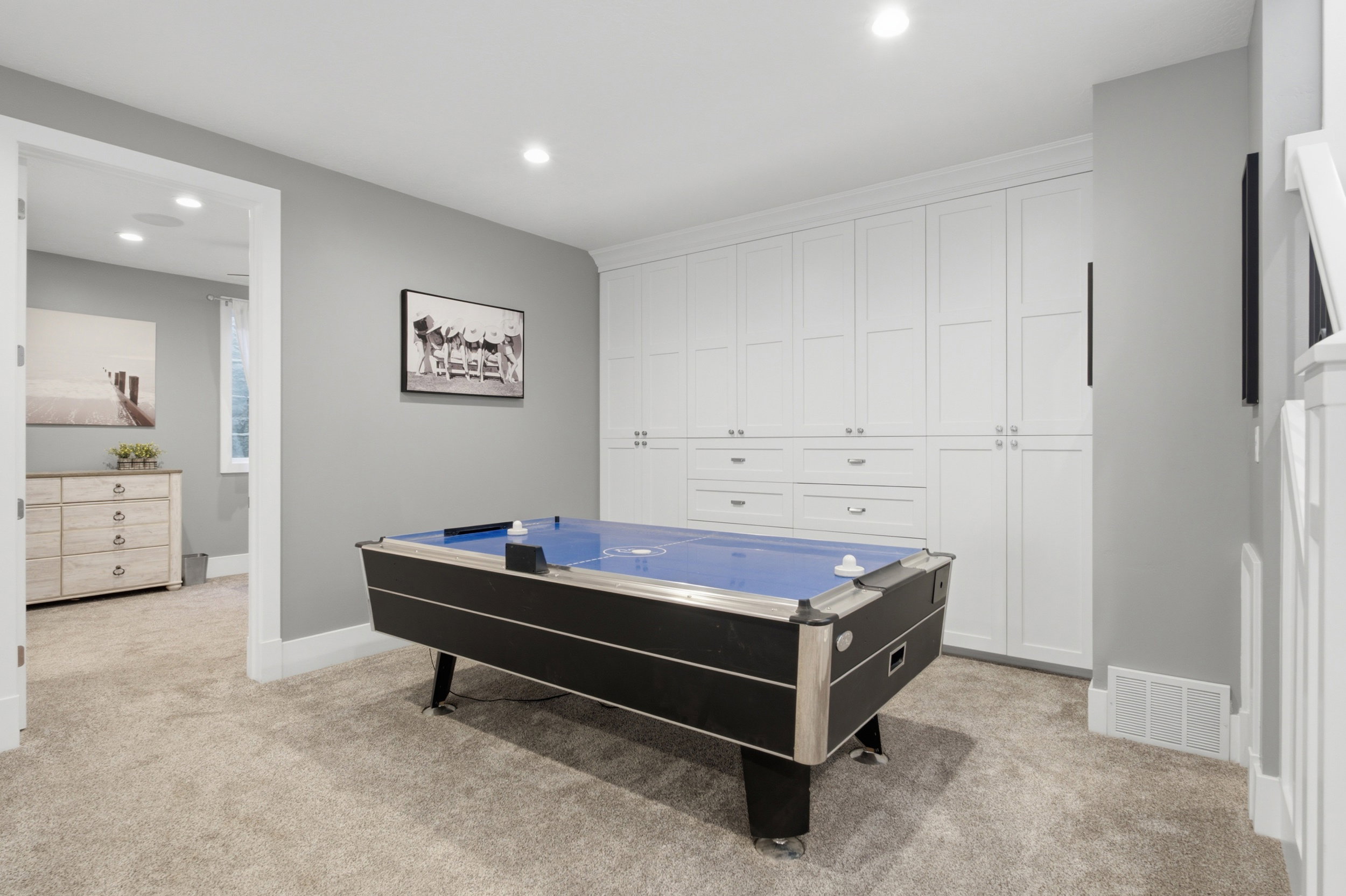 Recessed lighting illuminating a spacious room with light gray walls and plush carpeting, featuring extensive built-in white cabinetry with drawers and storage