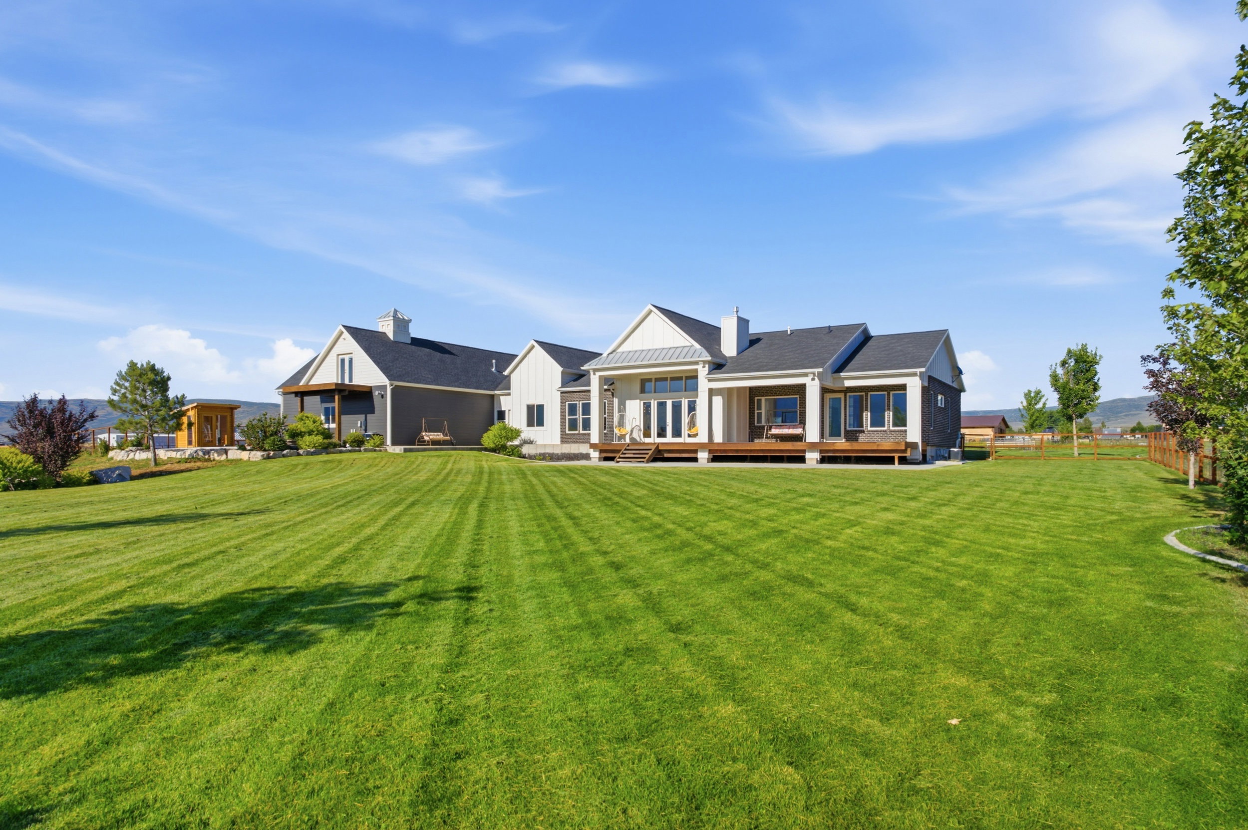 Expansive green lawn with a contemporary multi-gabled home featuring a covered porch, outdoor decking, multiple windows, and a partially fenced perimeter