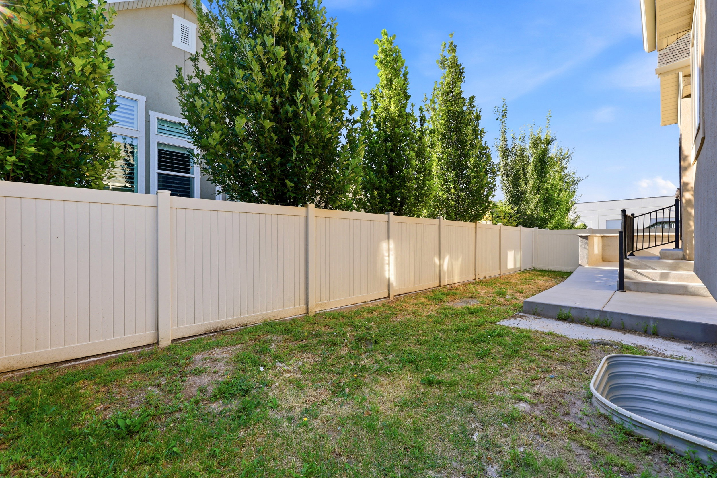 Enclosed backyard featuring a light-toned privacy fence, established trees, a concrete patio area, and exterior steps with a black metal railing